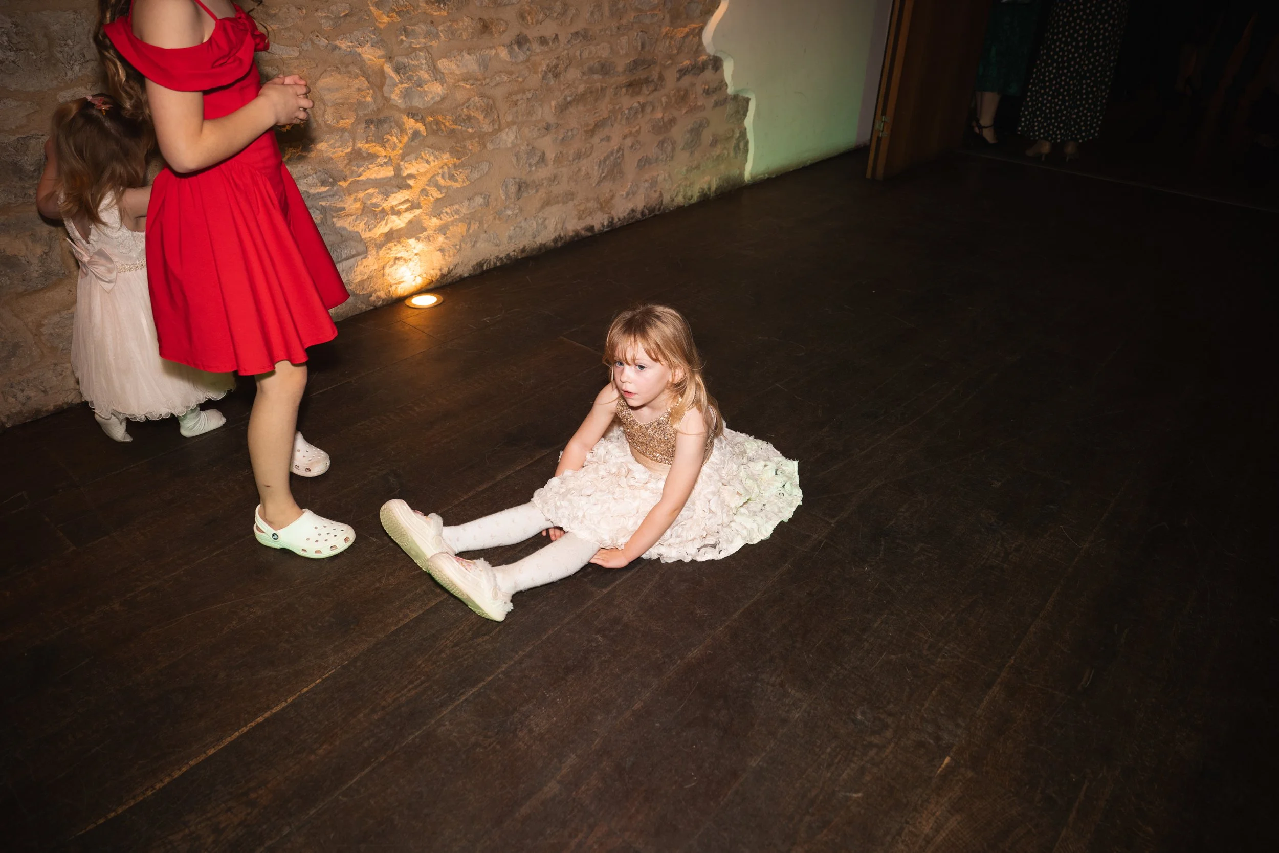 A young girl in a white dress with a gold top is sitting on the dark wooden floor, looking distressed. Two other girls in dresses, one in red and the other in white, stand nearby against a brick wall. The setting suggests an indoor event or party.
