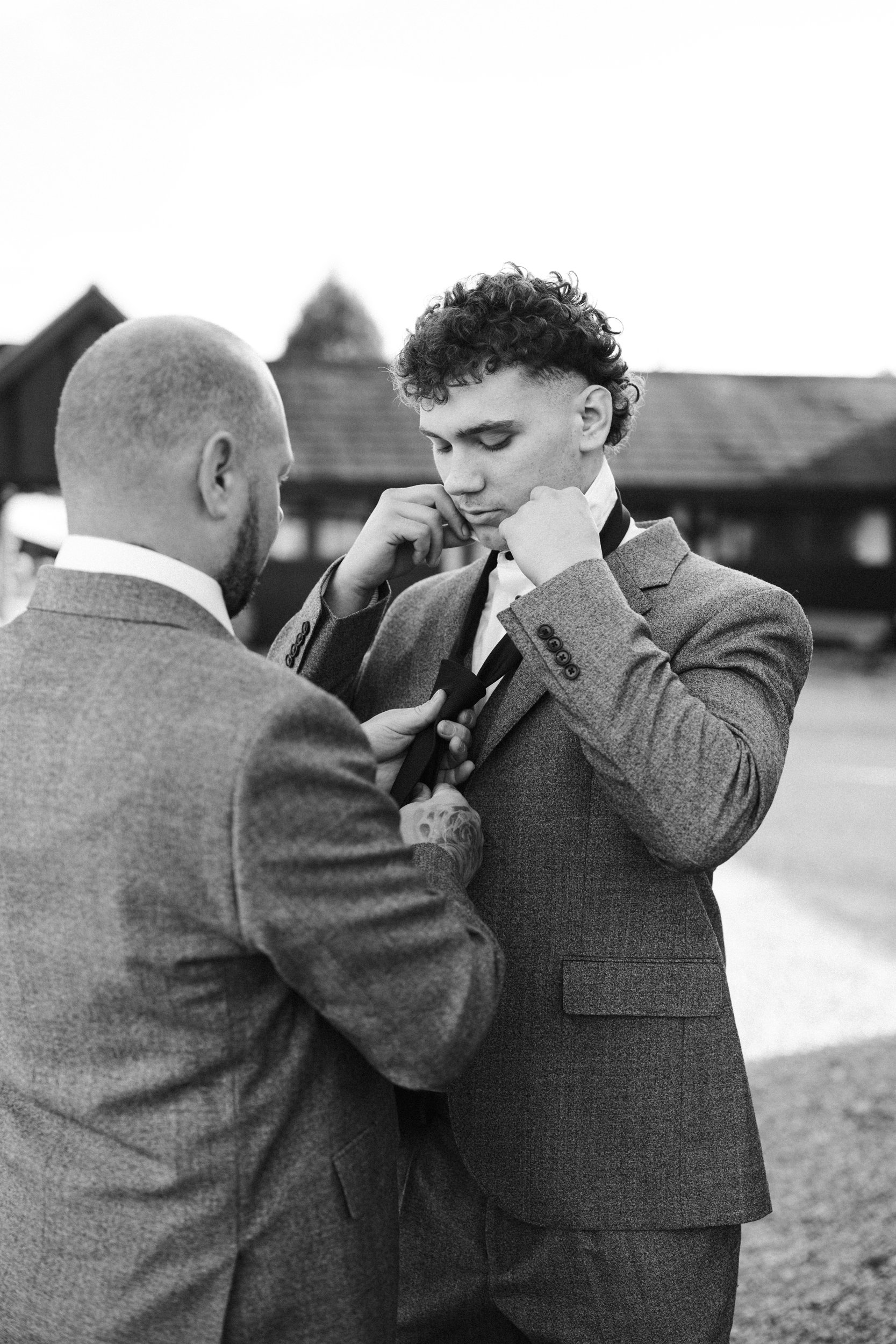 Two men in suits preparing for a formal event outdoors, with one adjusting the other's tie.