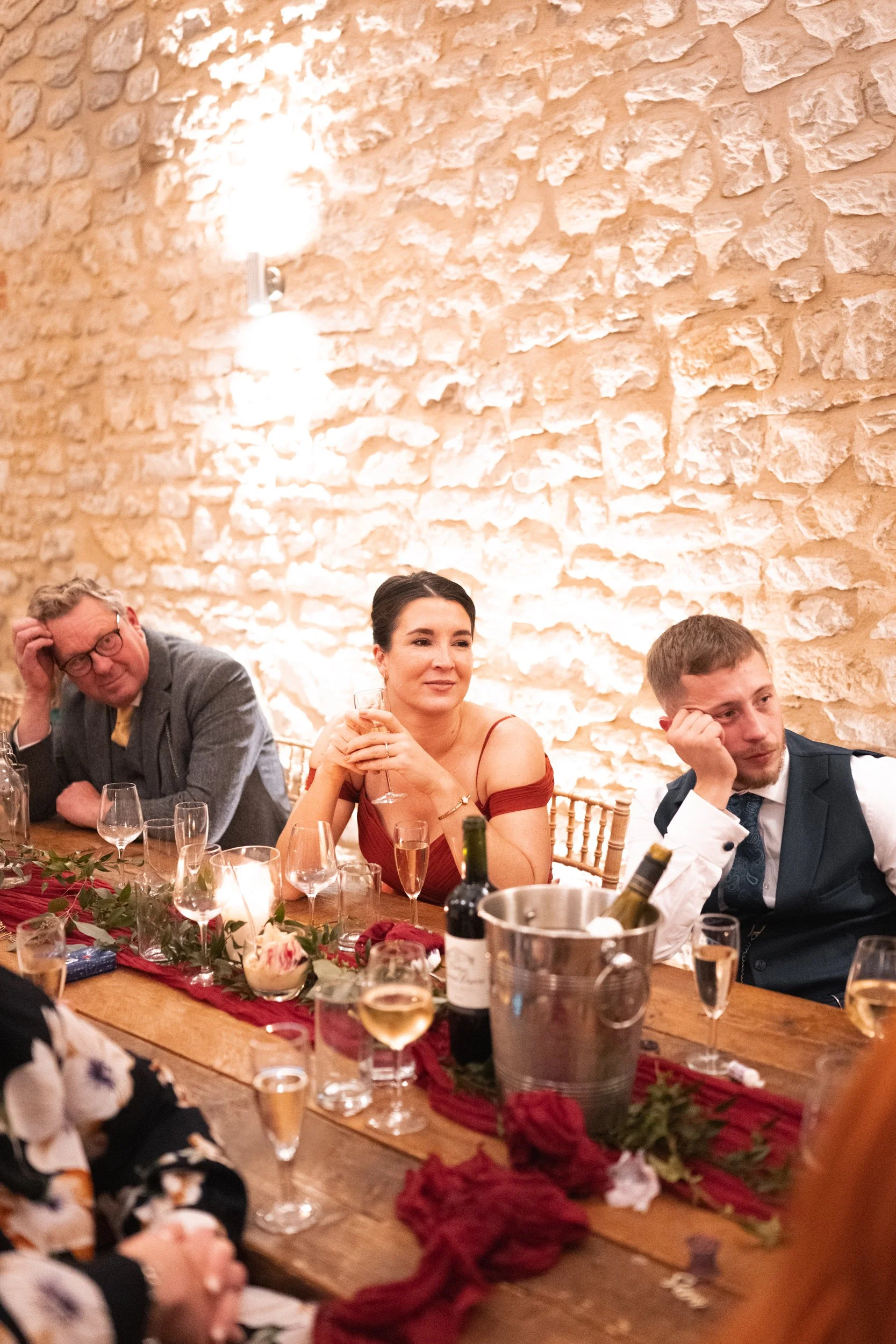 People sitting at a decorated dinner table with wine bottles and glasses, against a stone wall, at what appears to be a formal celebration or wedding reception.
