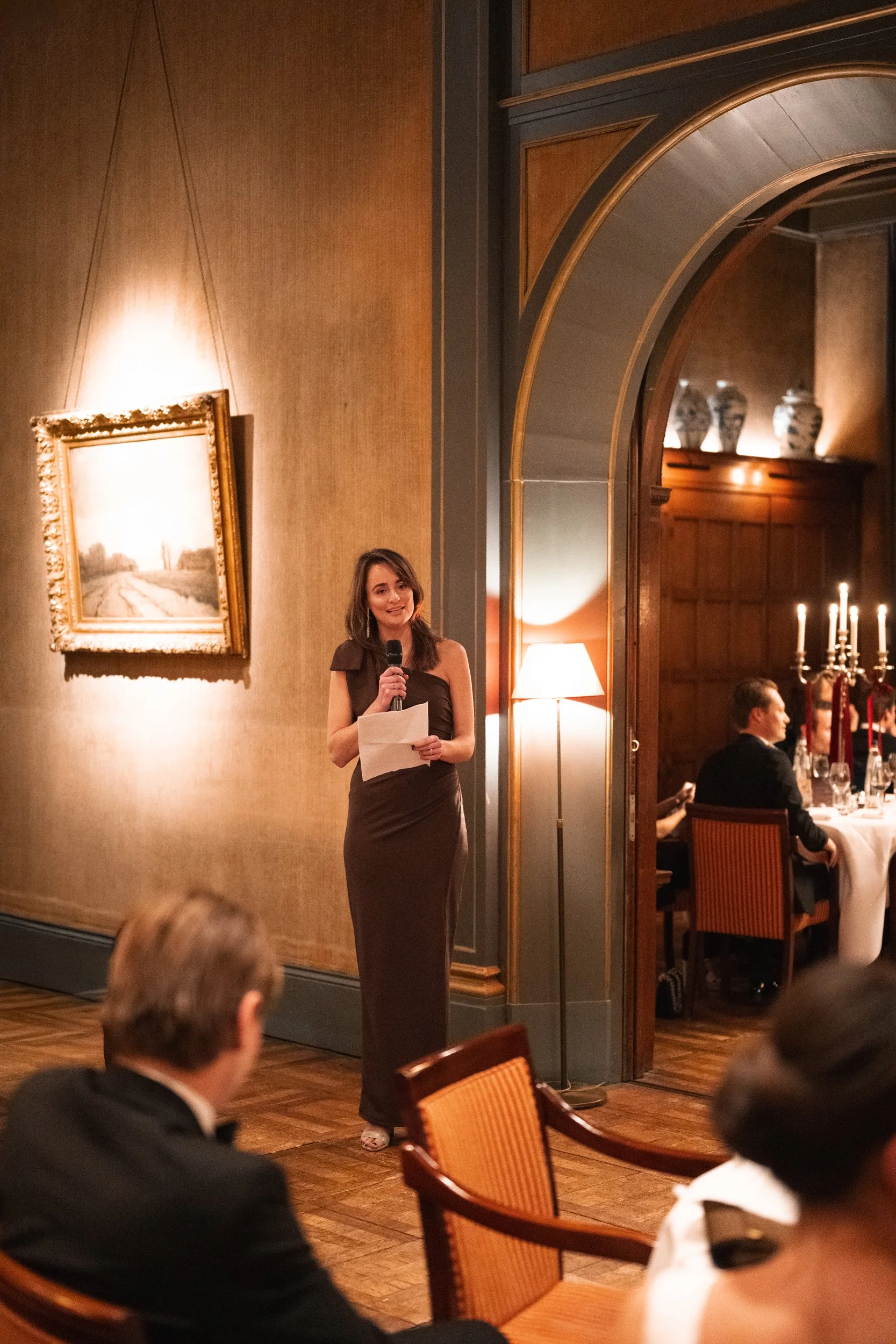 A woman in a dark dress giving a speech at a formal event in an elegant room with warm lighting and traditional decor.