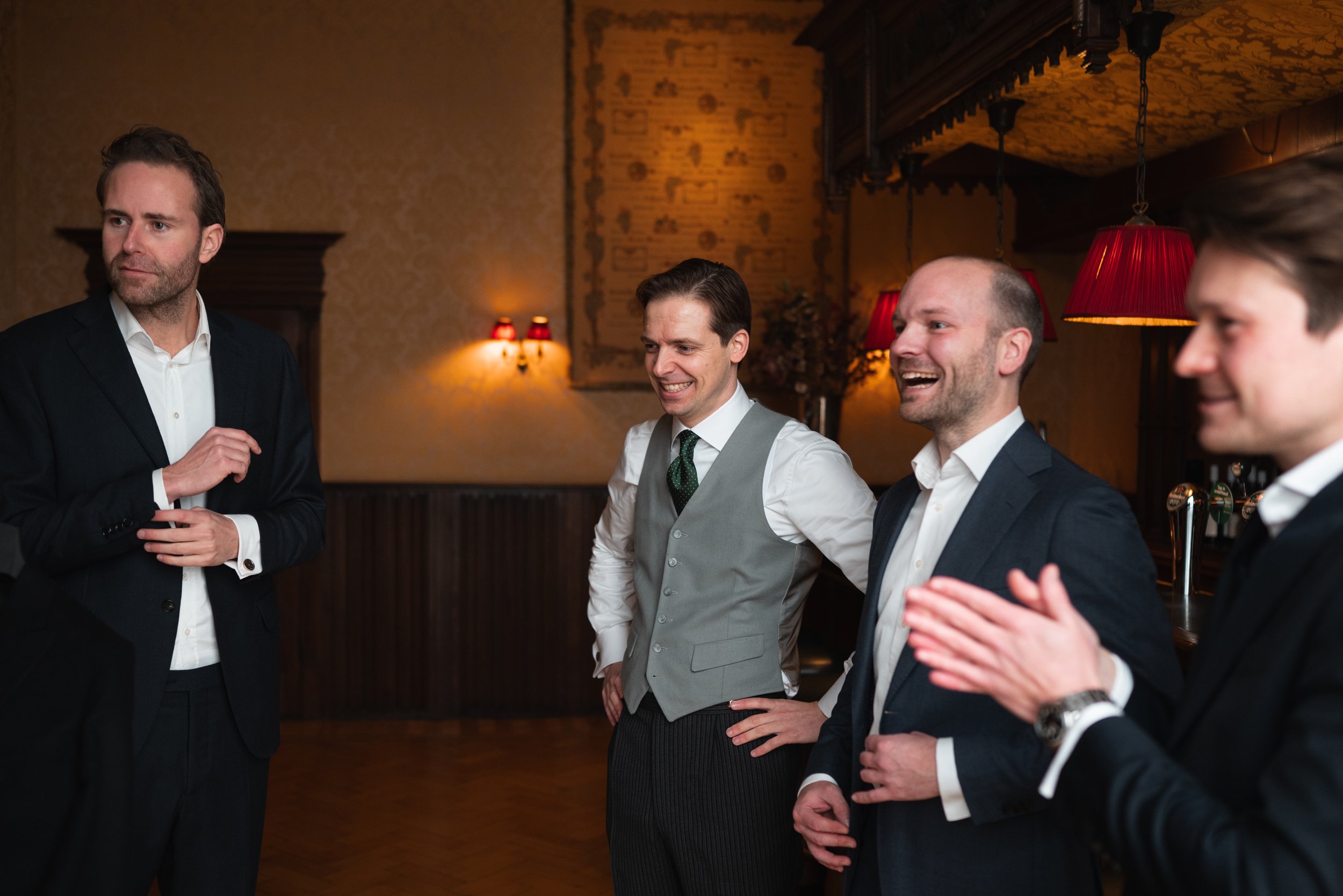 Group of four men in suits at a social gathering, smiling and clapping in a warmly lit room with red lampshades and vintage decor.