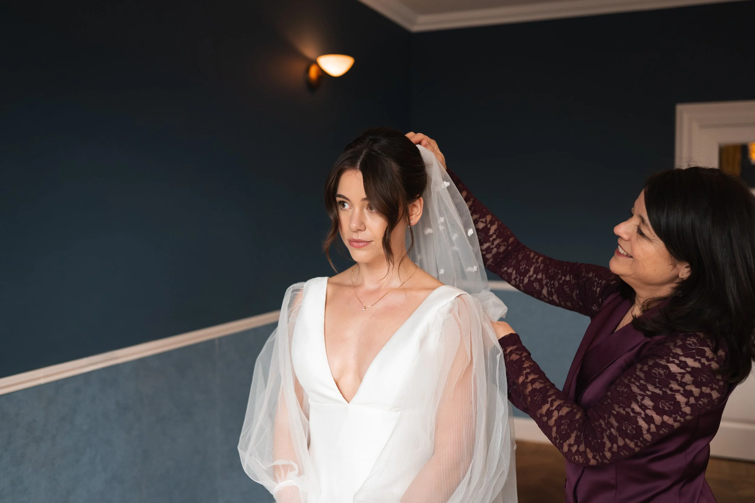 A woman in a white wedding dress with sheer sleeves is having a veil adjusted by another woman in a burgundy lace dress, in a room with dark blue walls.