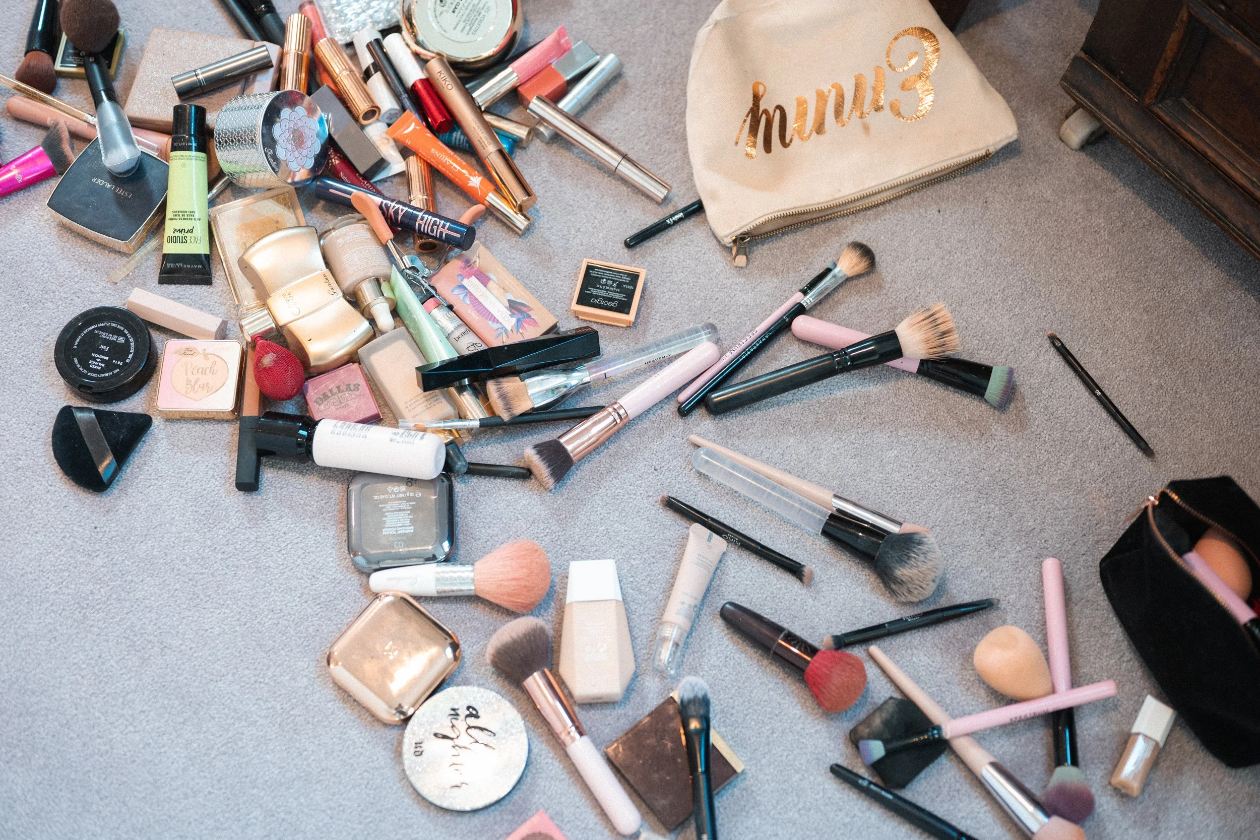 An assortment of makeup products and brushes scattered on a carpeted floor, with a beige cosmetic bag with 'mumu3' written in gold, and a black travel makeup bag.