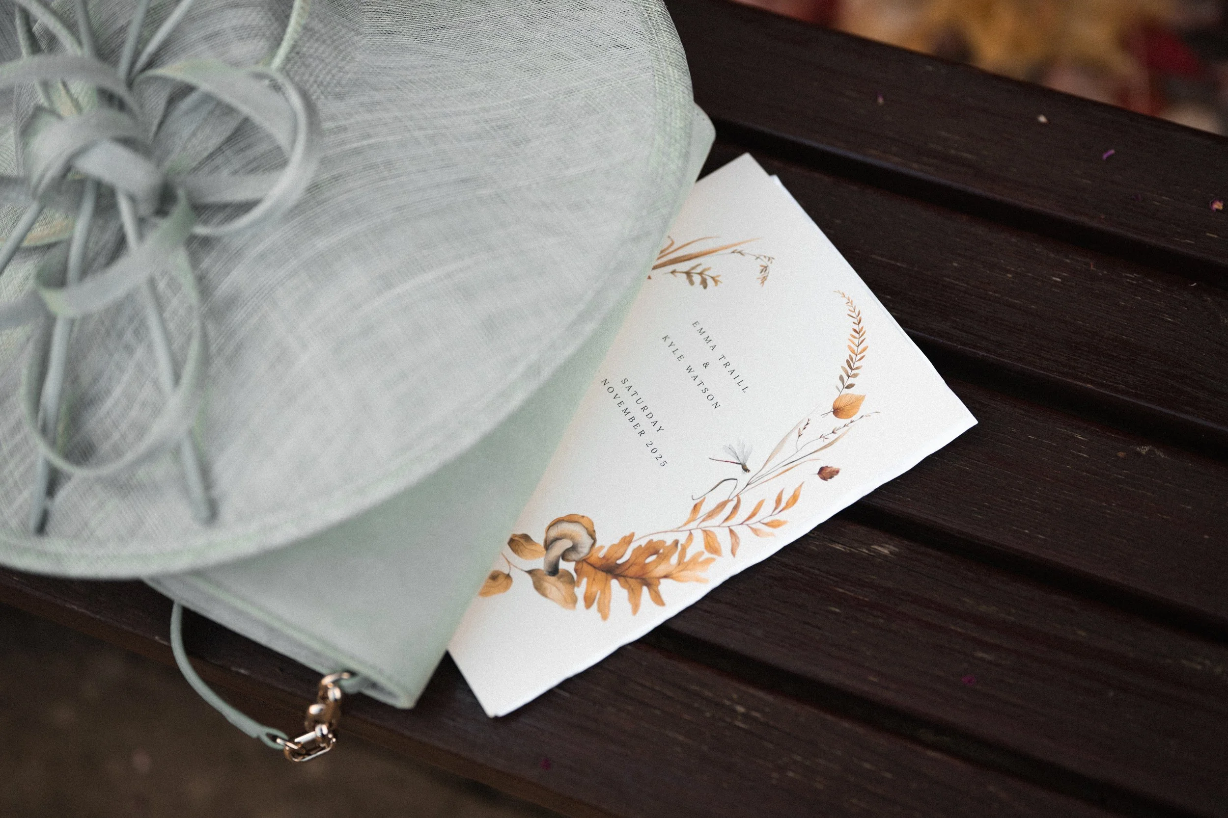 A white wedding invitation with botanical illustrations is on a dark wooden bench, partially covered by a pastel green fascinator hat with a bow and netting.