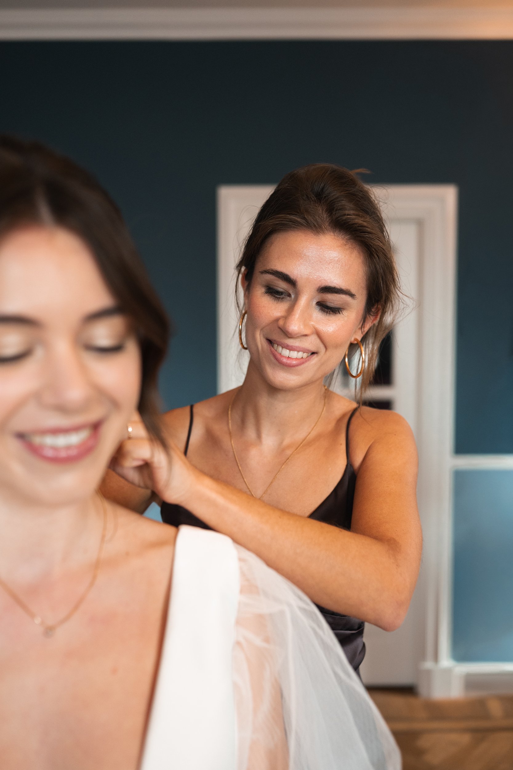 Two women smiling, one helping the other with her wedding dress or gown, in a room with dark blue walls and white doors.