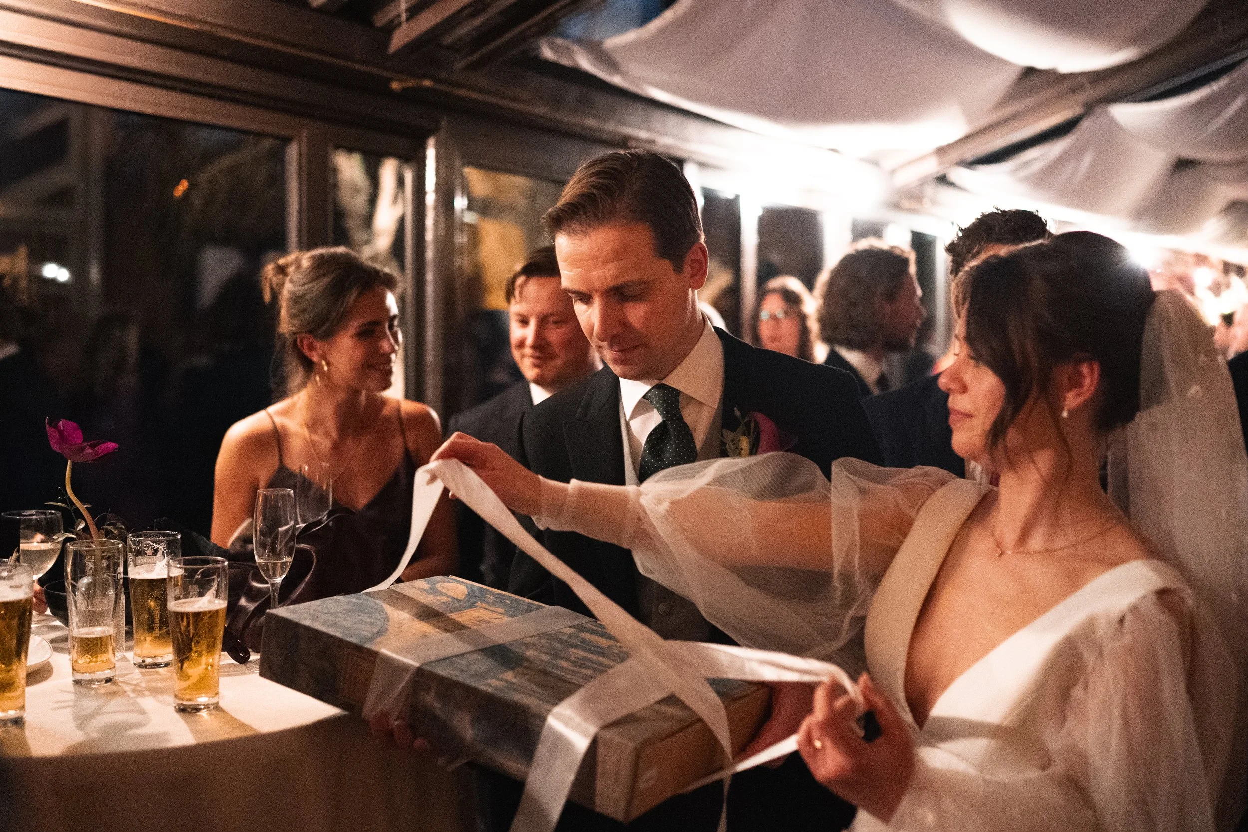 A wedding reception with guests dressed in formal attire, with a man and woman exchanging a gift, and others in the background.