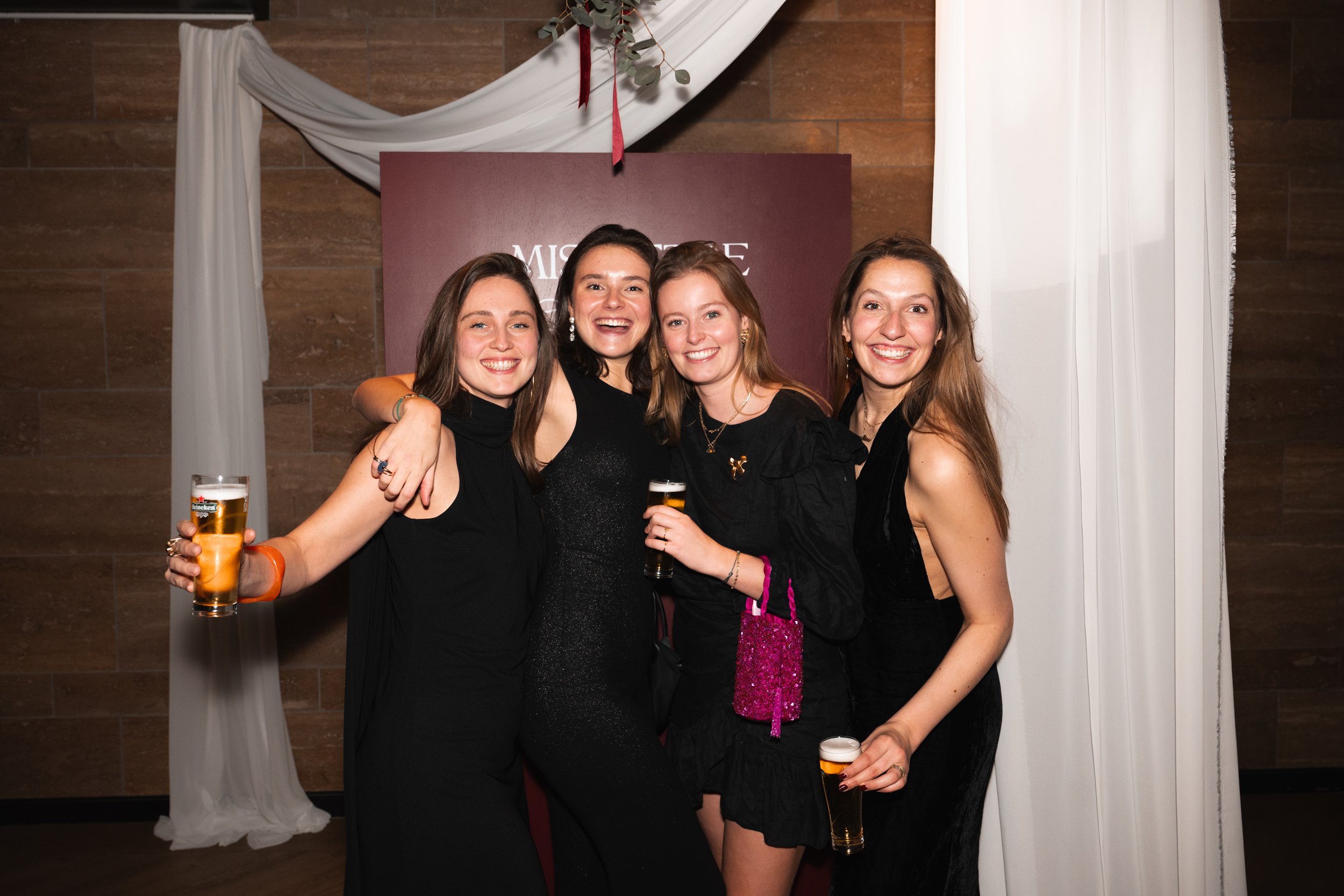 Four women smiling and posing with drinks at a celebration or party, standing in front of a maroon background with white drapery.