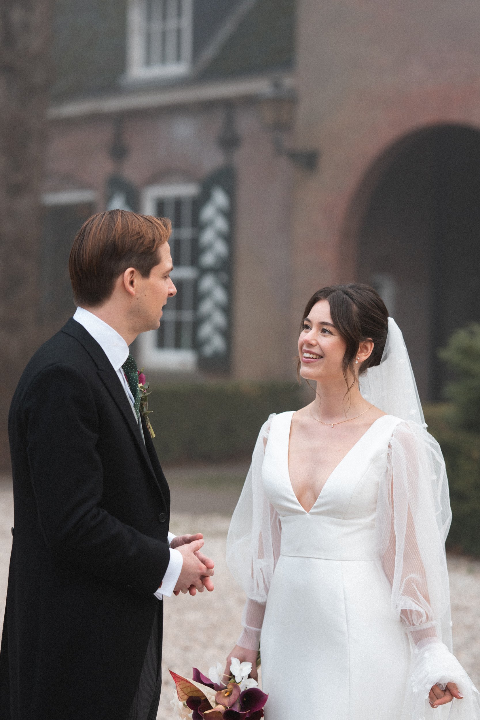 A man in a black suit and a woman in a white wedding dress standing outdoors, smiling at each other, with a blurred building in the background.