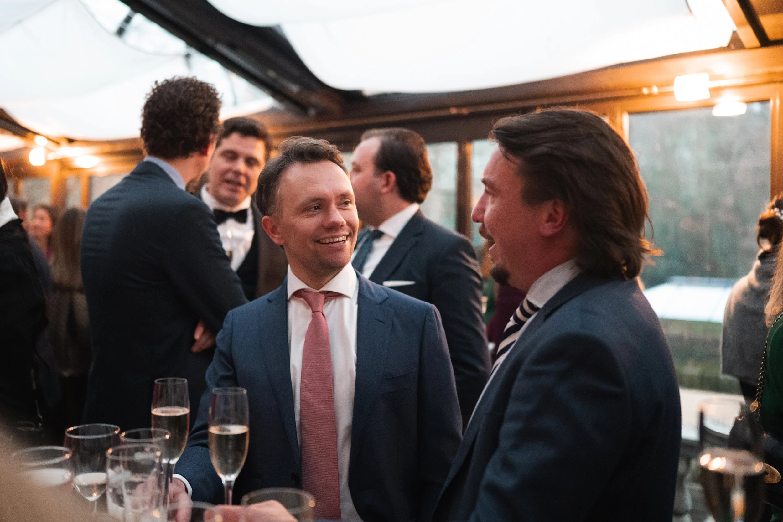 Group of well-dressed men engaging in conversation at a social event or party, with glasses of champagne on the table in a warmly lit, enclosed space.