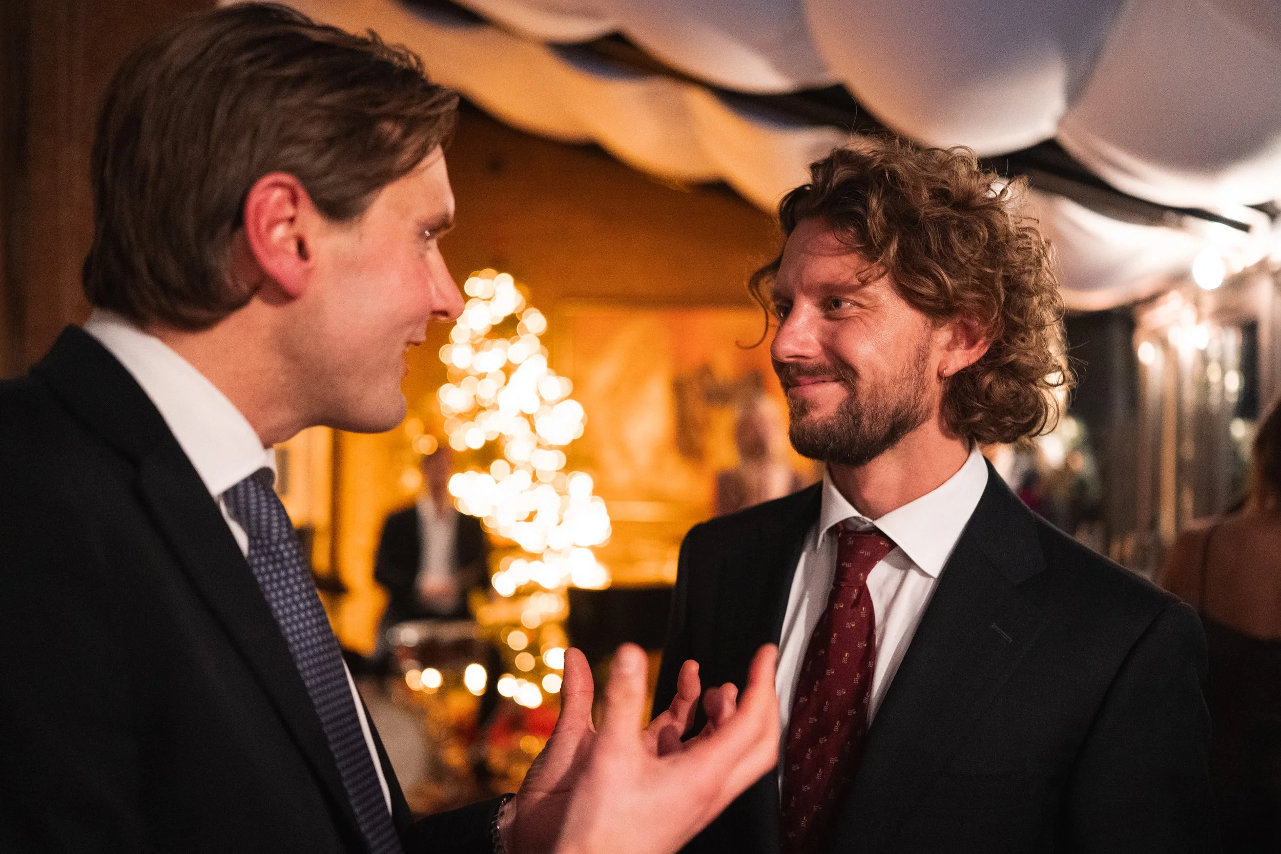Two men in suits engaged in a friendly conversation at a festive indoor event, with warm lighting and Christmas decorations in the background.