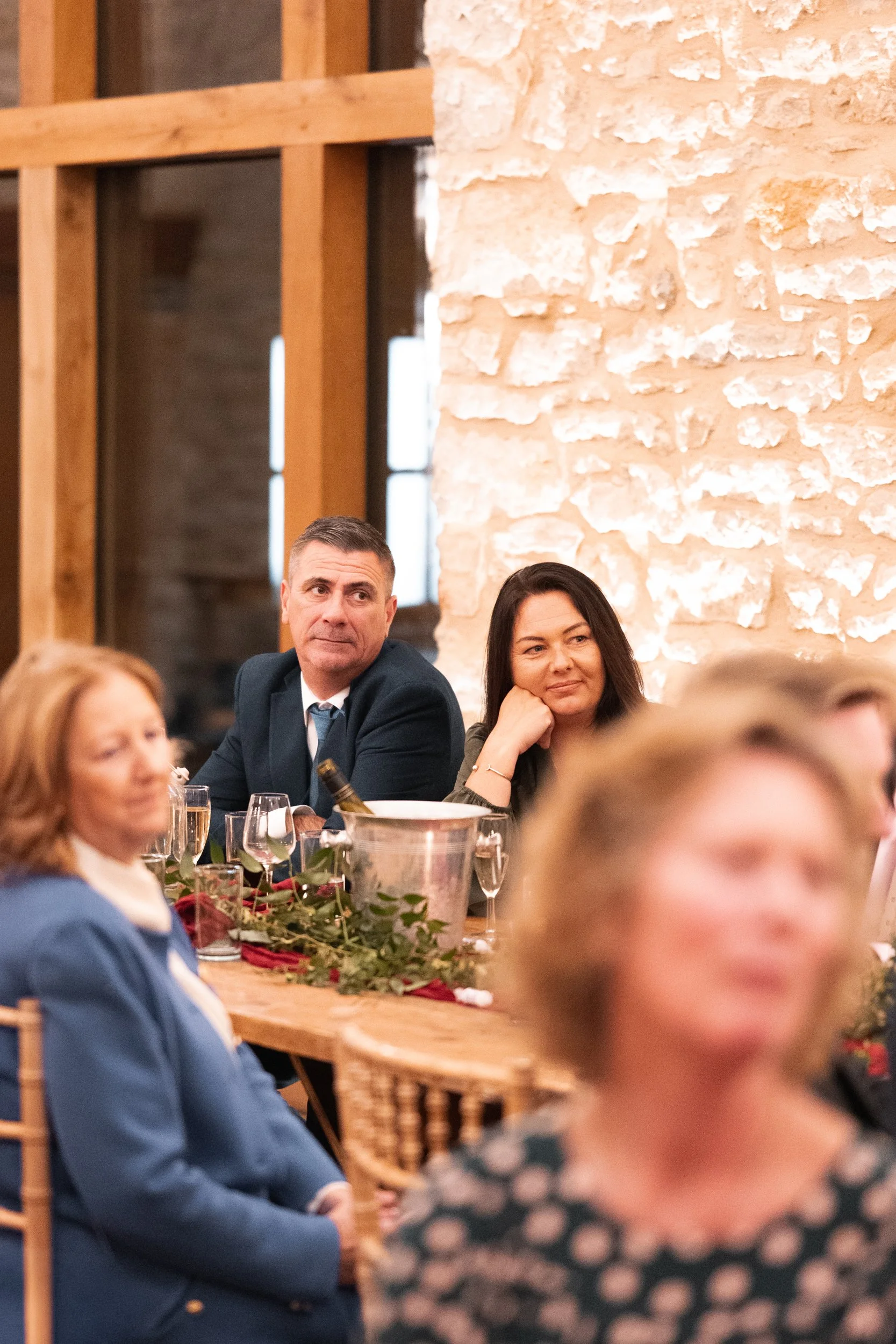 People sitting at a decorated table during a formal gathering, with two women and a man prominently visible, one woman is blurry and a woman with dark hair is resting her head on her hand.