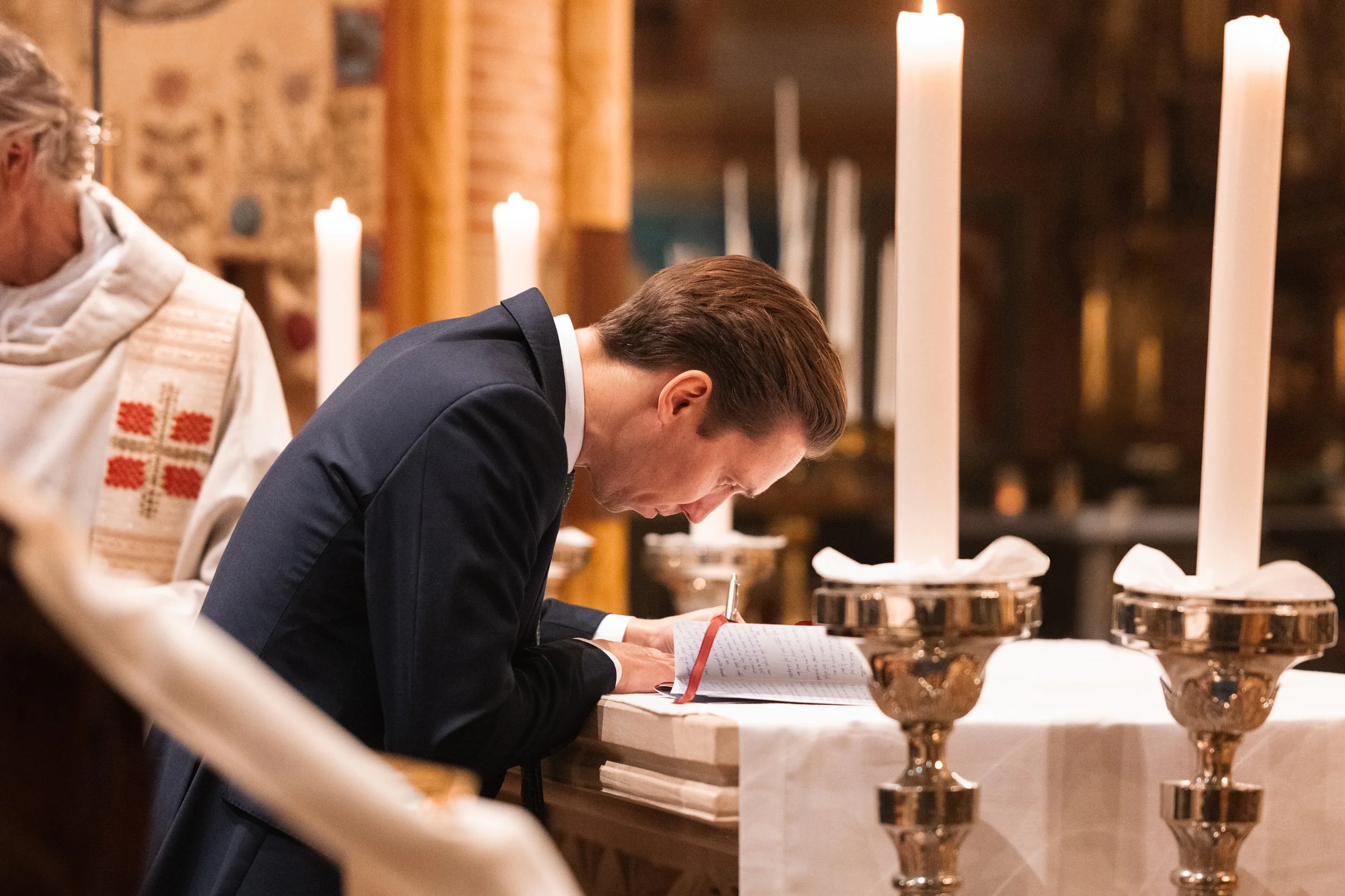 A man in a dark suit leaning over a prayer book on a church altar, with tall lit candles and a woman in religious attire nearby.