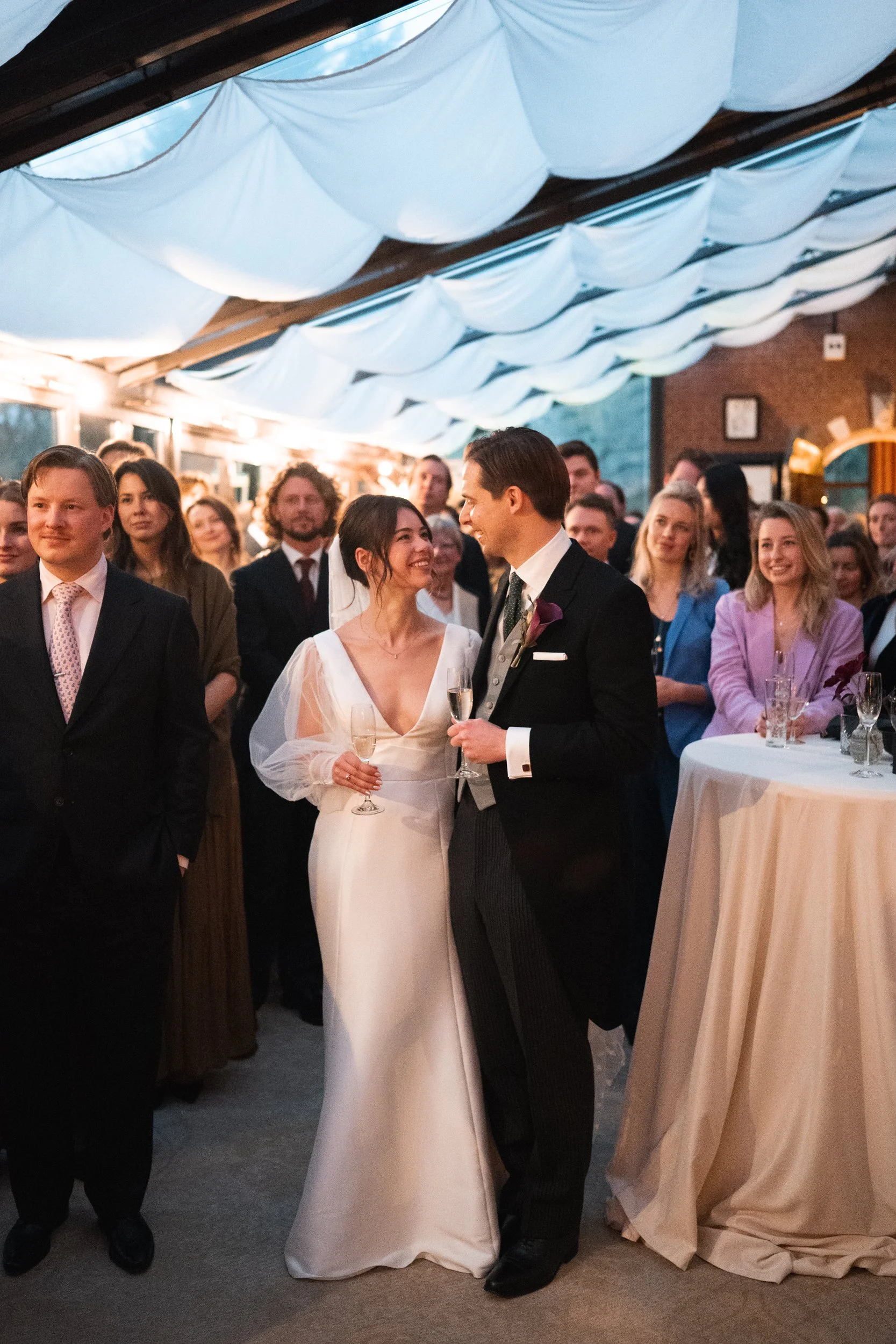 A bride and groom at a wedding reception, holding champagne glasses and smiling at each other surrounded by guests.