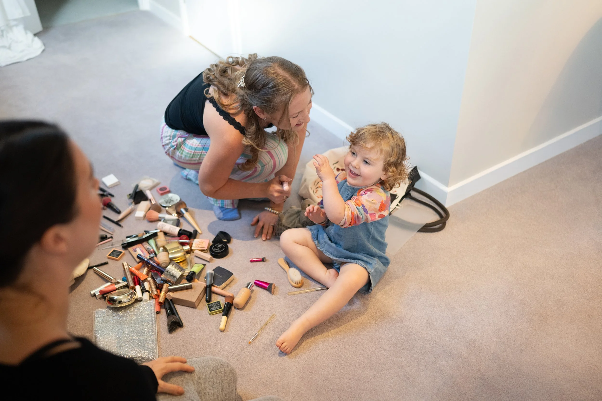 A young girl with curly hair and a denim dress sitting on the floor, smiling and playful, surrounded by scattered makeup products. An adult woman with long hair and a black top is kneeling nearby, engaged in conversation with her. Another woman with 