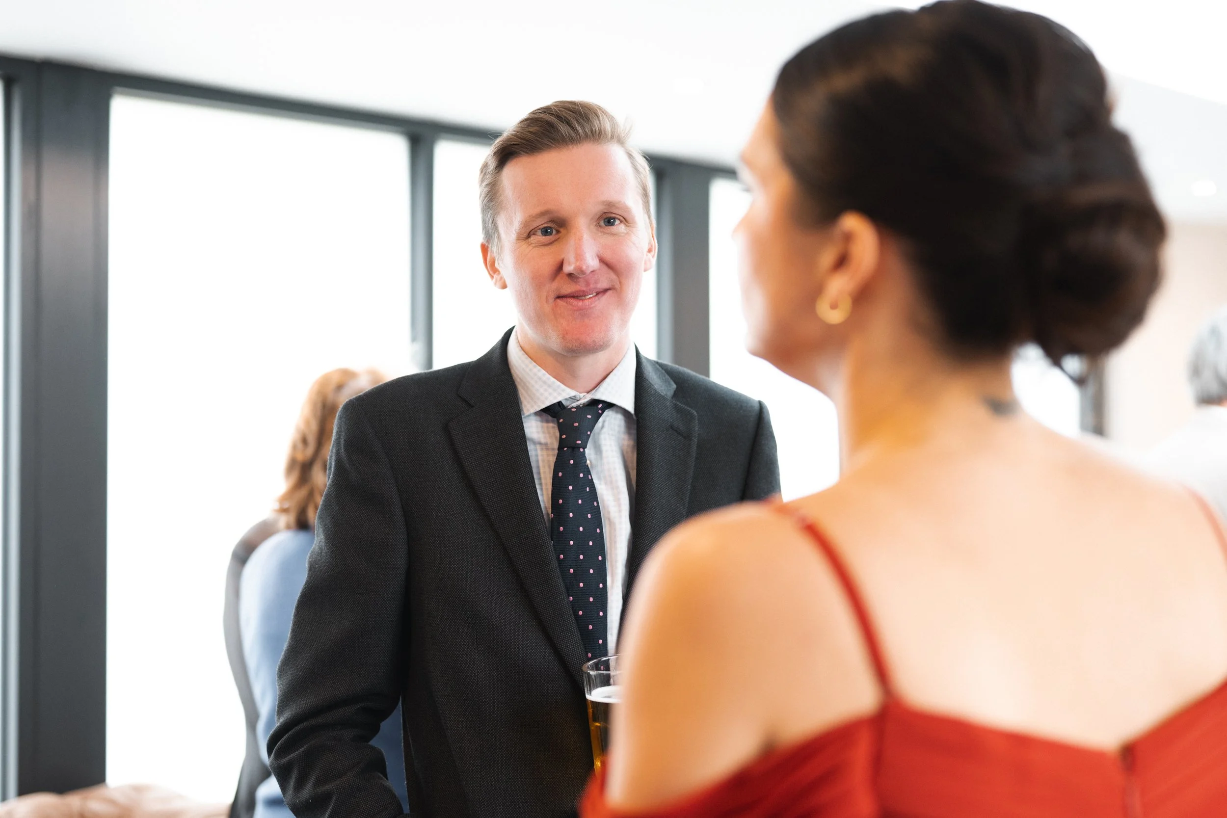 A man in a suit holding a drink and smiling, talking to a woman in an off-shoulder red dress at a social event.