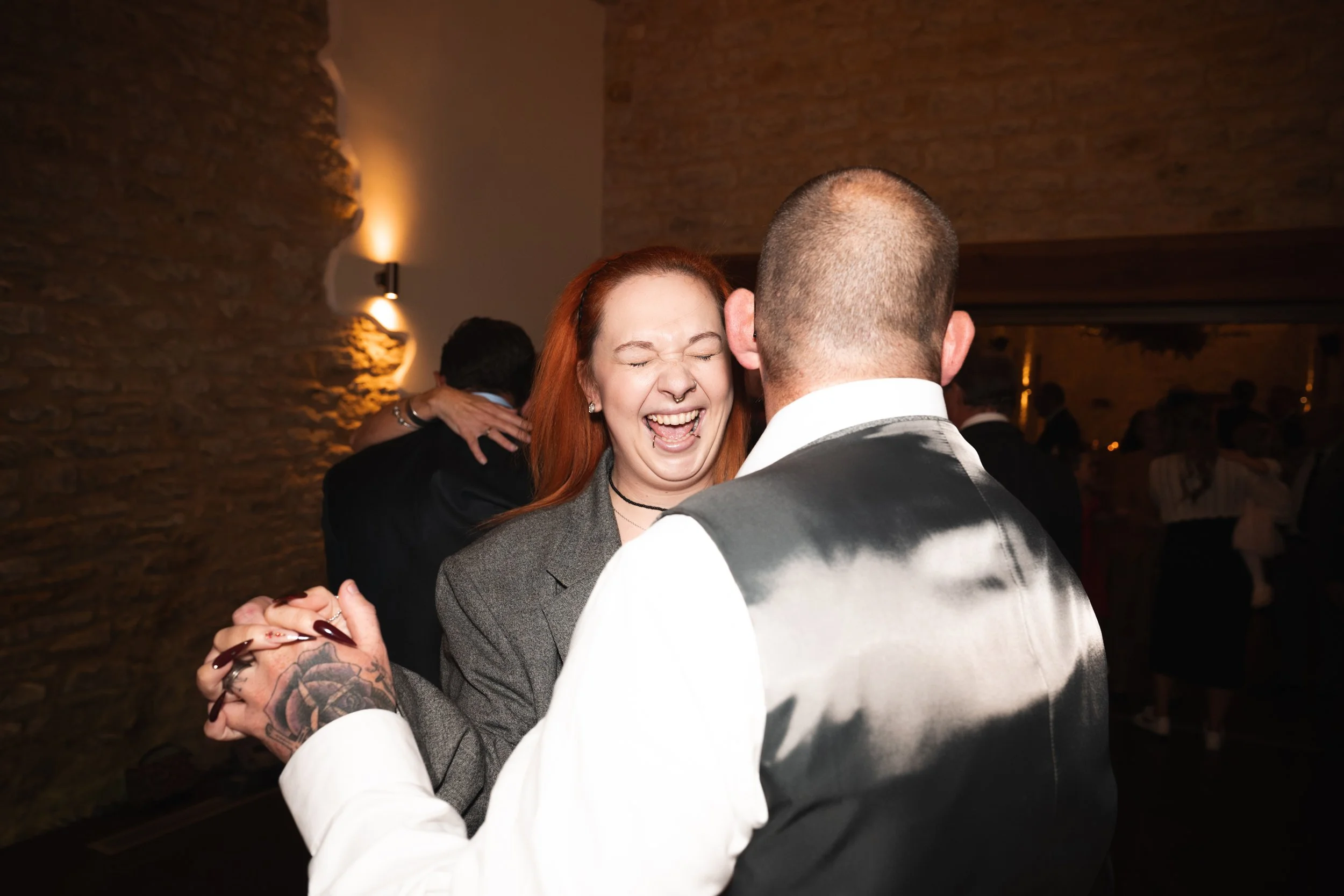 A woman with red hair and a man with a tattooed hand are dancing and smiling at a social event in a dimly lit room with brick walls.