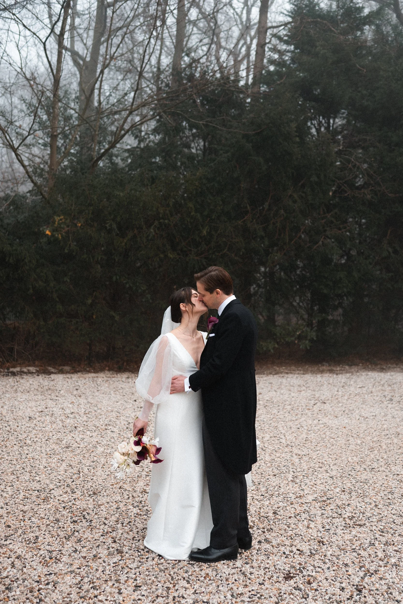 A bride and groom kissing outdoors on their wedding day, holding a bouquet of flowers, with trees in the background.