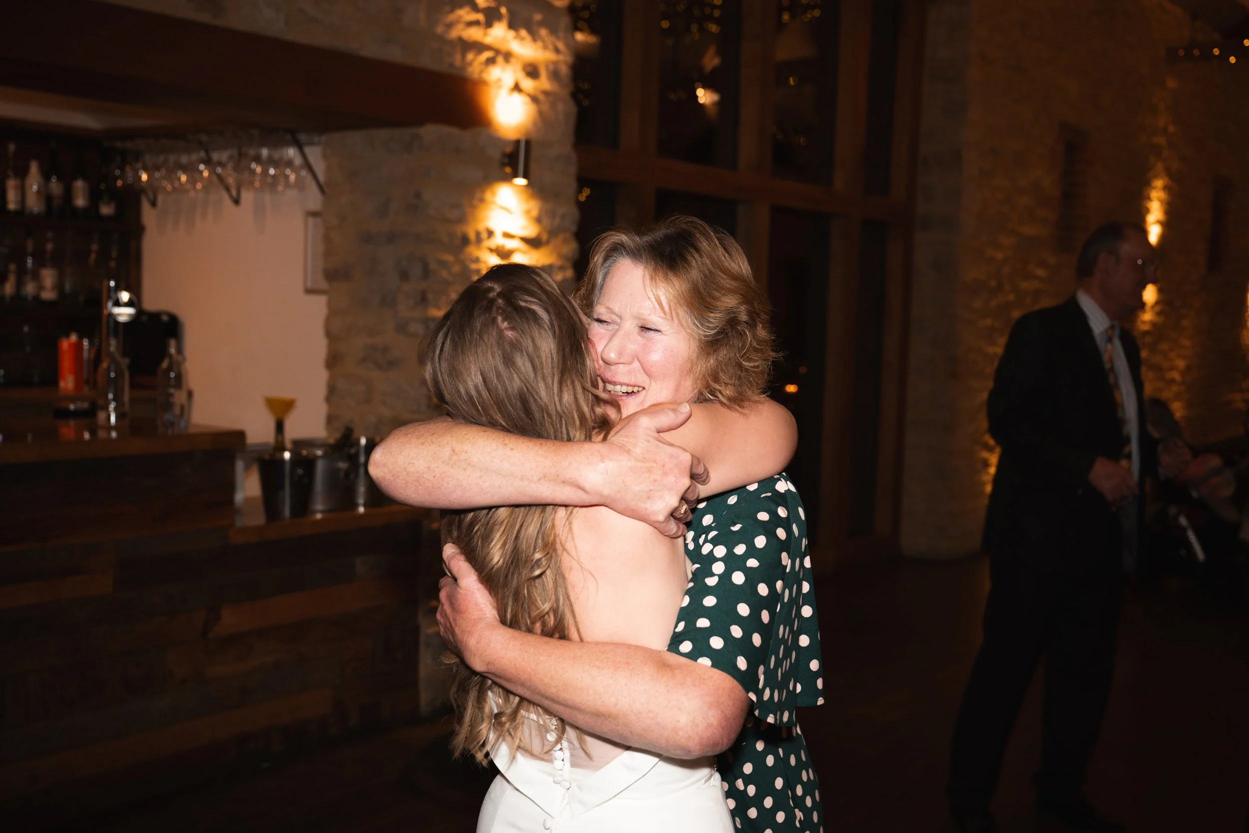 A woman and a girl are hugging and smiling at each other at a party or celebration, with a bar and a man in the background.