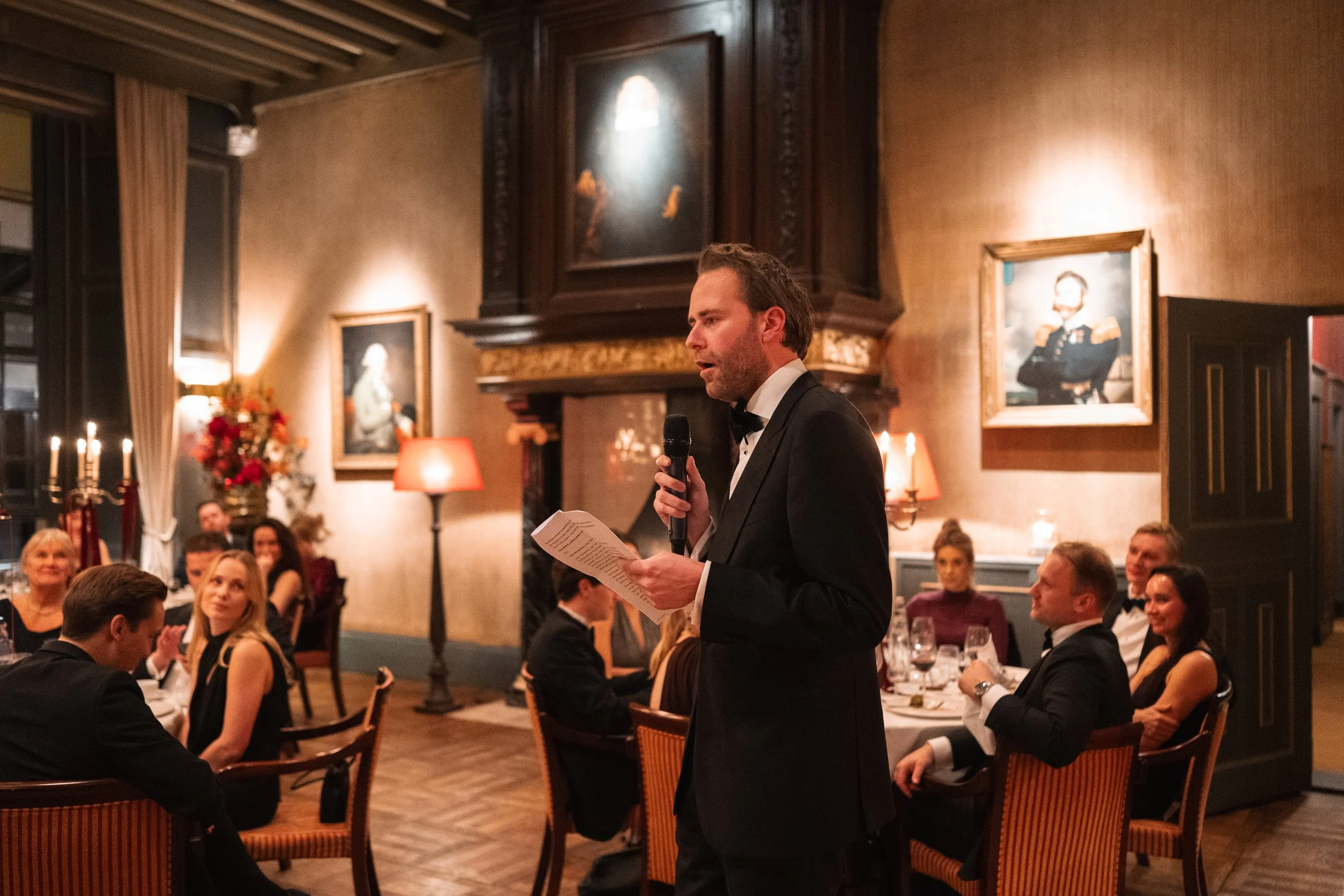 A man in a tuxedo giving a speech at a formal dinner event in an elegant room with guests seated at tables, framed portraits on the walls, and dim lighting.