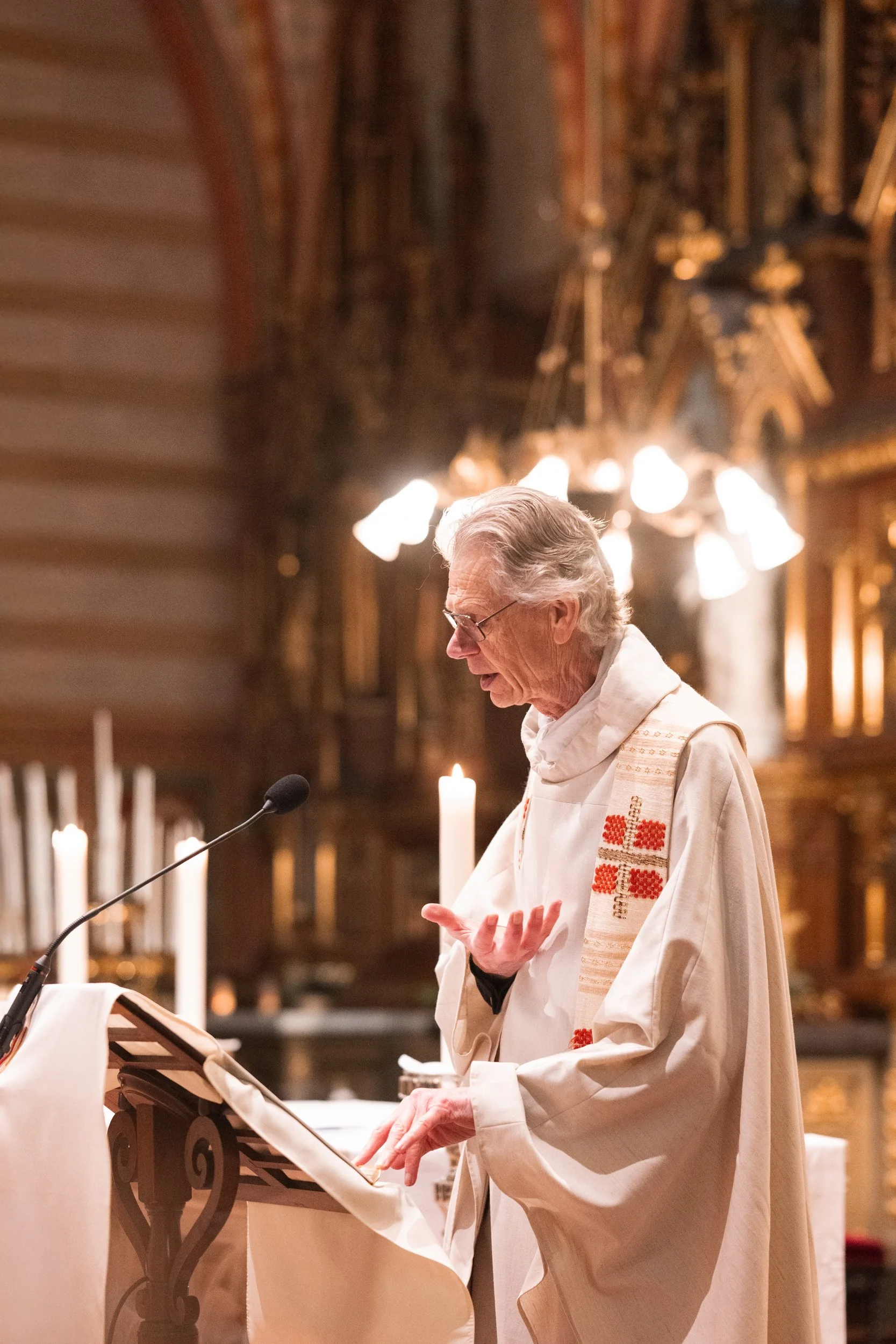 An elderly man in religious robes reading or speaking at a pulpit in a church with lit candles and ornate woodwork in the background.