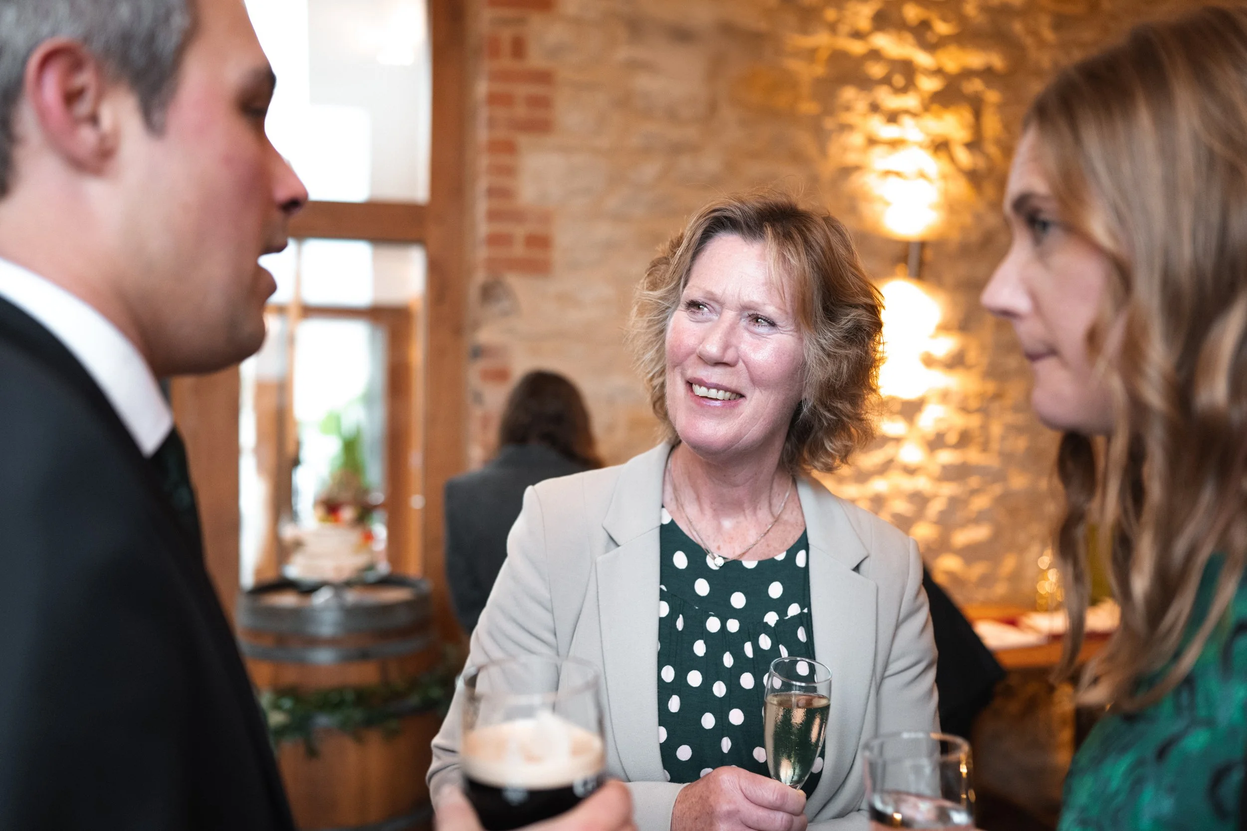 Three people at a social gathering are engaged in conversation, with one woman holding a glass of champagne and a man holding a glass of dark beer. The middle woman is smiling and listening to the individuals on her sides, in a warmly lit room with b