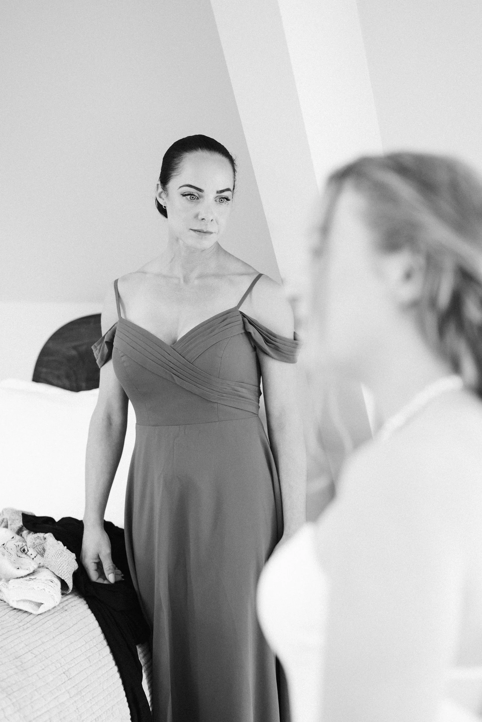Black and white photo of a woman in an elegant off-shoulder dress looking at her reflection in a mirror.
