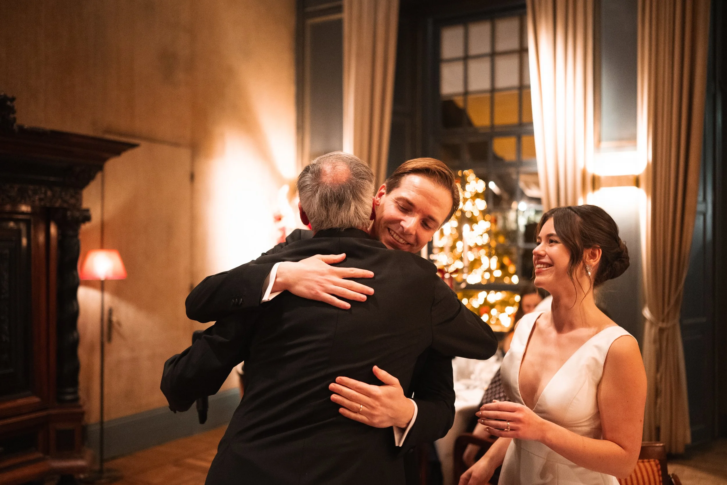 A man in a suit hugging an older man at a wedding reception, with a woman in a white dress smiling nearby. Christmas tree and warm lighting in the background.