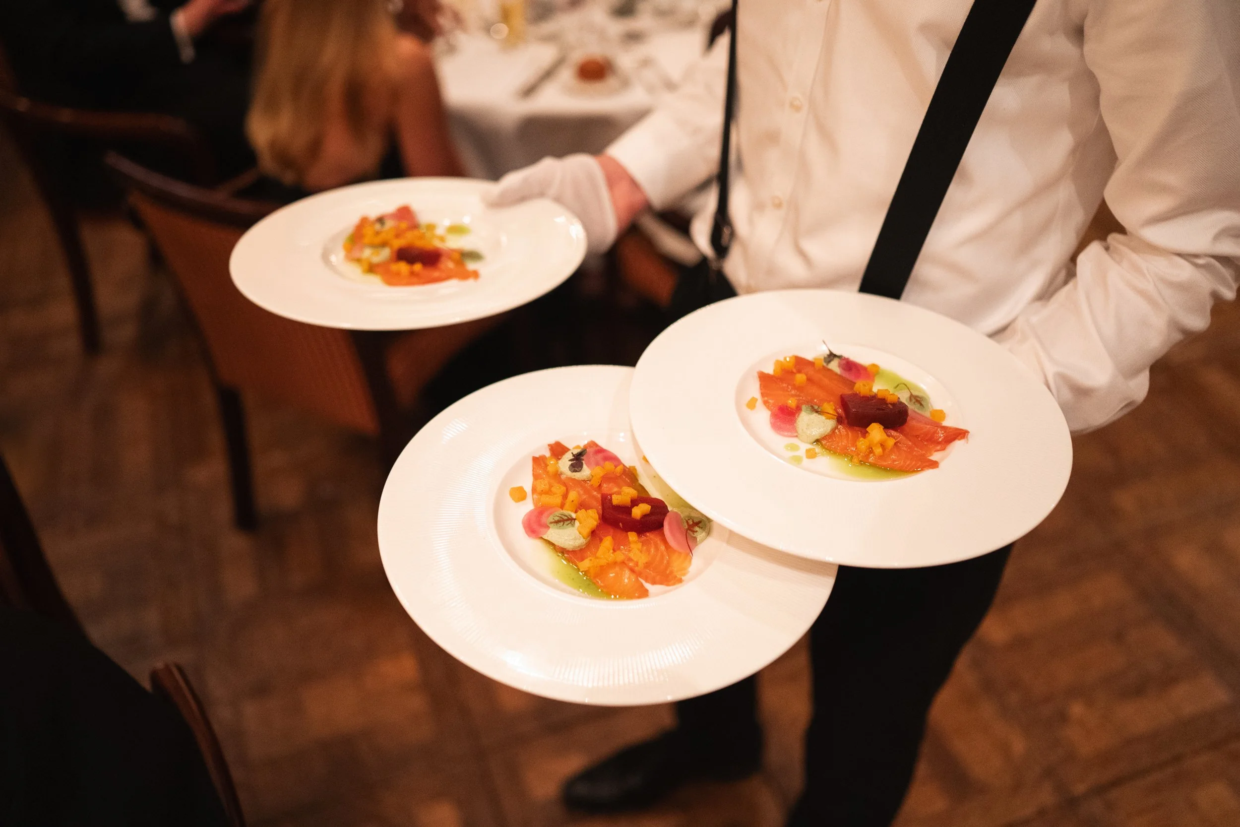A waiter holding three plates of colorful plated gourmet food in a restaurant.