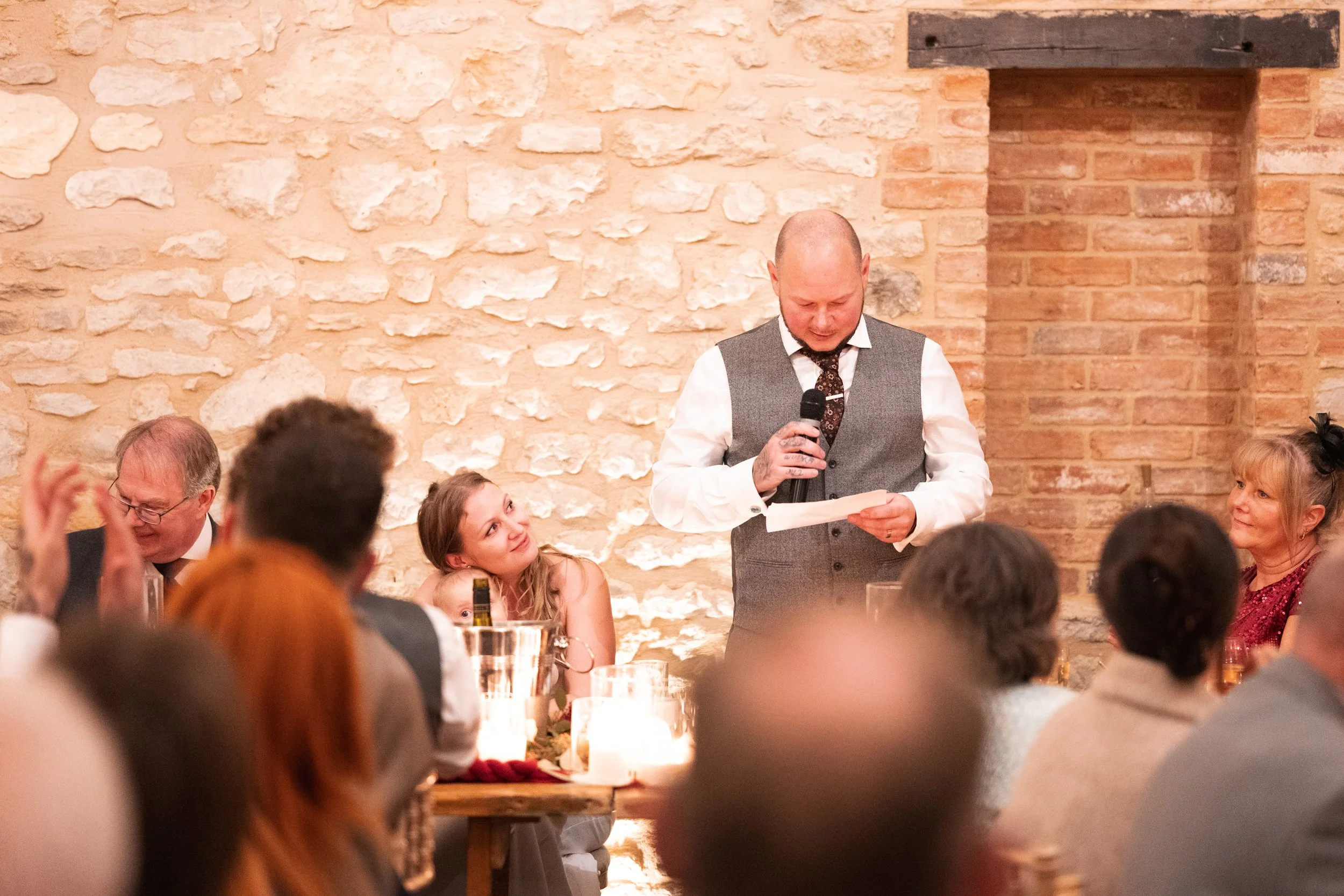 Man giving a speech at a wedding reception in a rustic brick room, surrounded by seated guests and candlelit tables.
