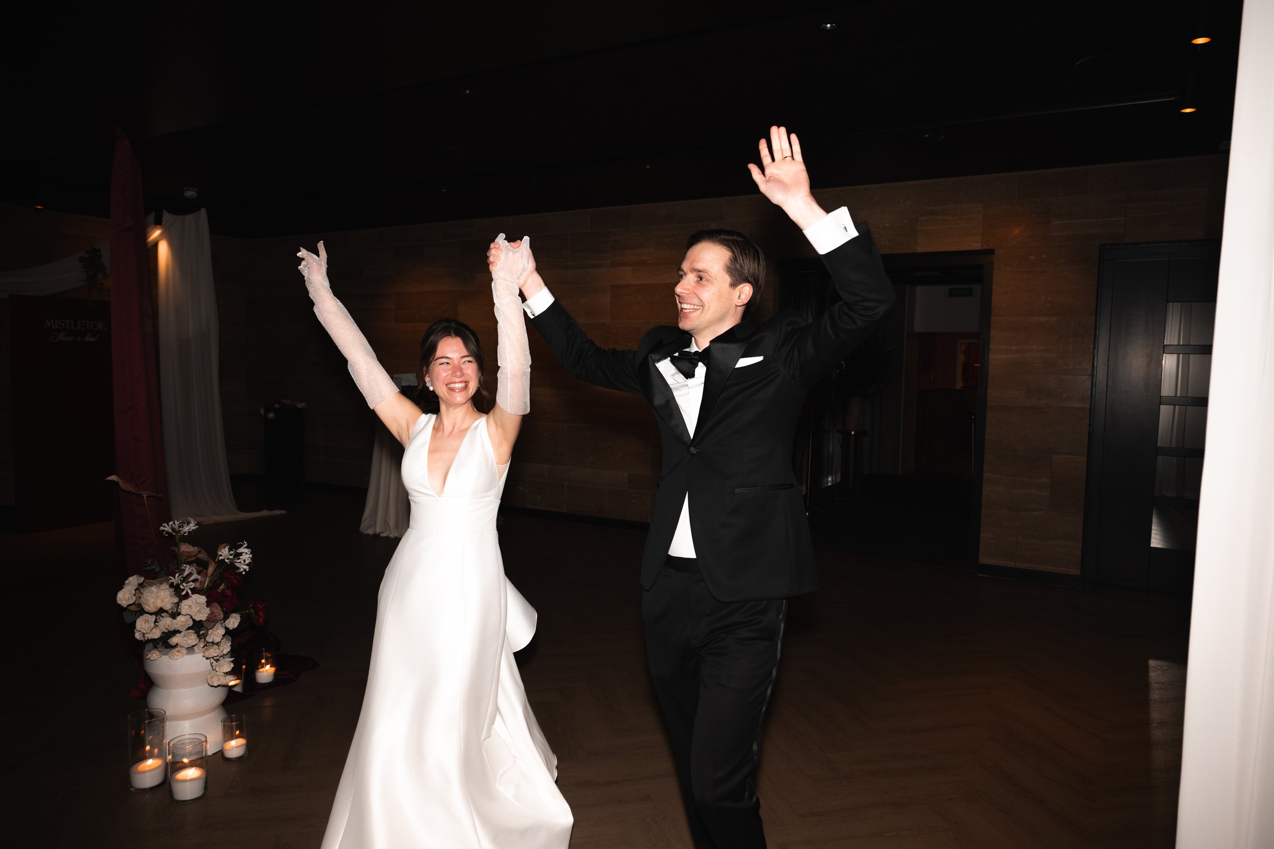 A bride and groom dancing at their wedding reception, smiling and holding hands, in a dimly lit venue with candles and floral decorations.