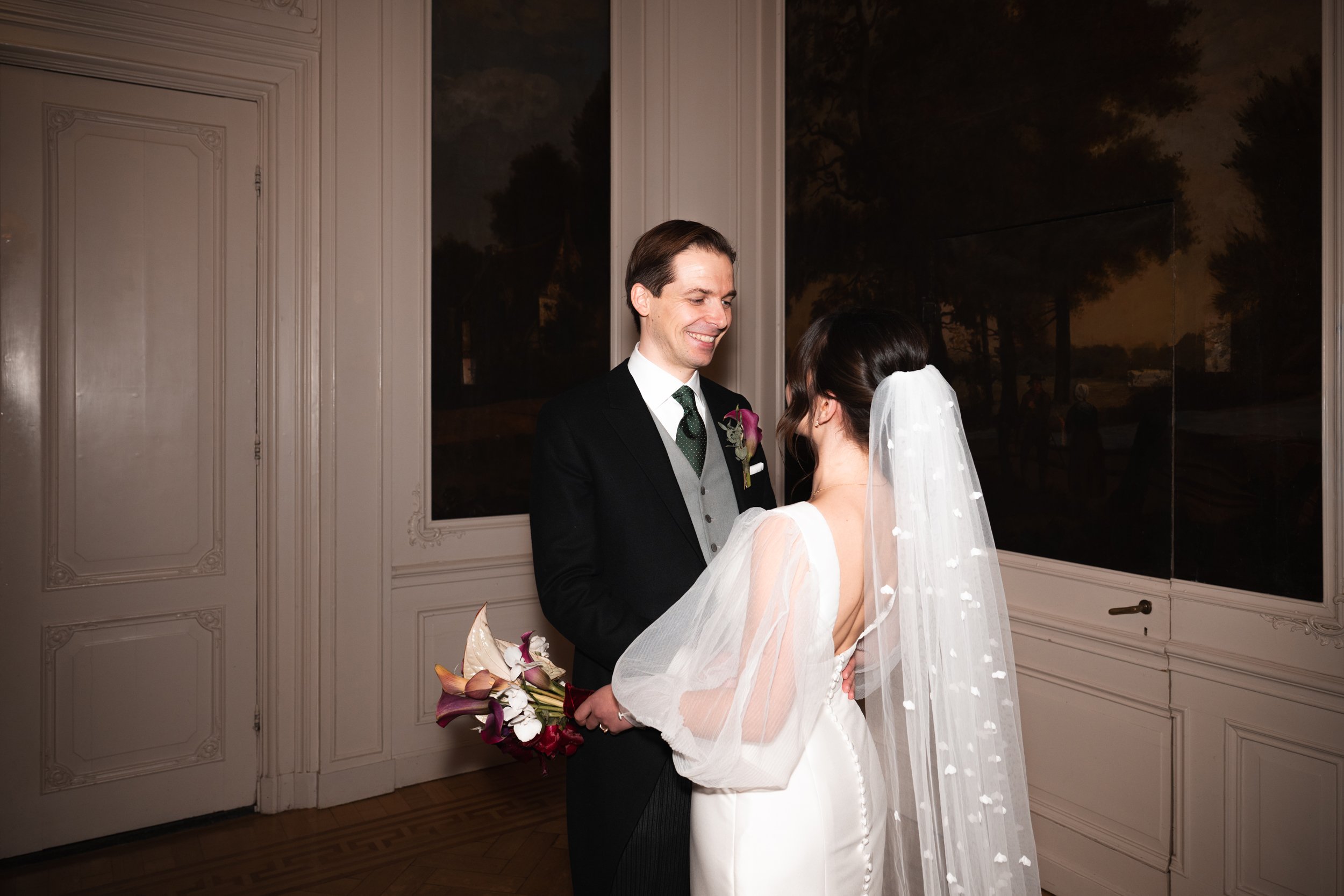 A bride and groom standing face to face during their wedding ceremony, indoors with classic white paneled walls and large windows showing a dark sky outside.