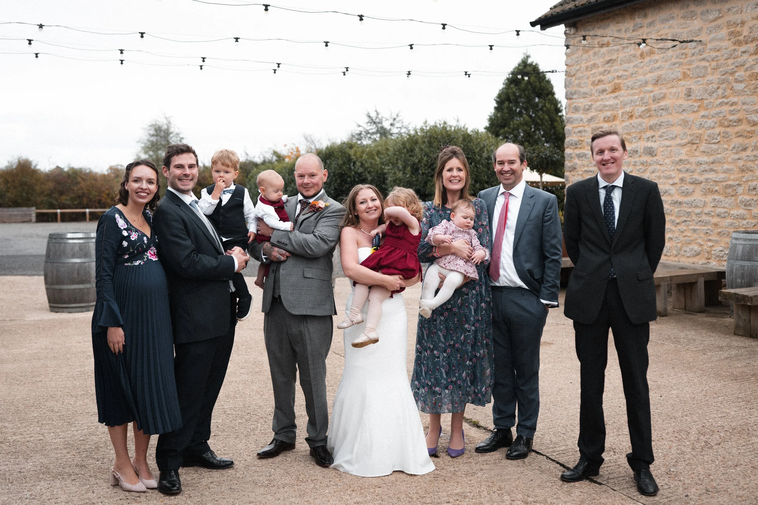 A group of nine people, including children, dressed in formal attire, standing outdoors near a stone building with string lights overhead, smiling for a photo at a wedding or special occasion.