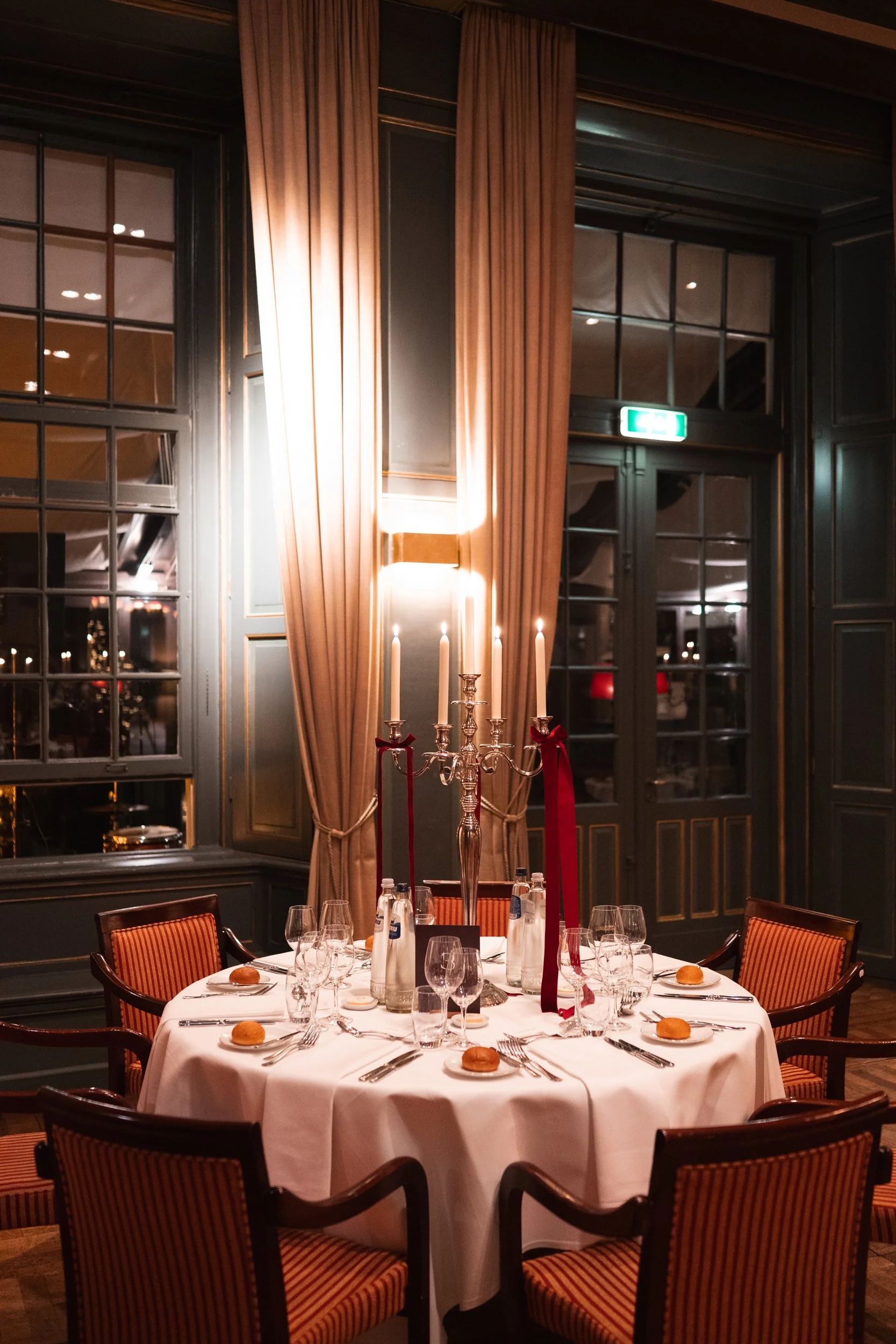 A round table set for dinner with white tablecloth, six glasses, silverware, and bread rolls. The centerpiece is a silver candelabrum with five lit candles decorated with red ribbons, in a dimly lit, elegant room with dark green walls, tall beige cur