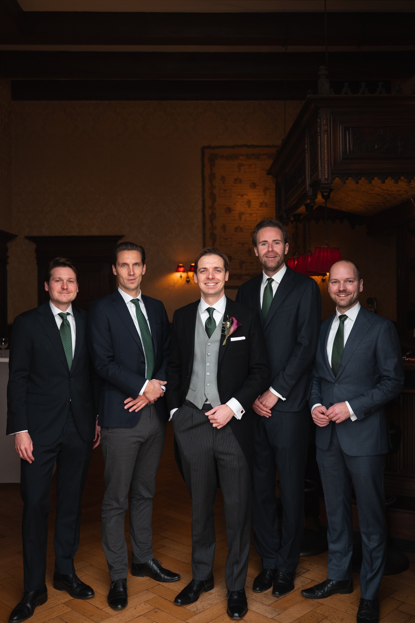 Group of five men dressed in suits, standing together indoors at a formal event, with dark wood furniture and subdued lighting in the background.
