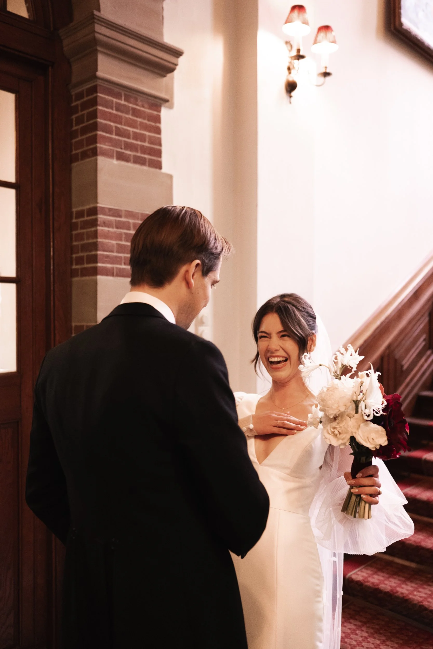 A bride and groom sharing a joyful moment inside a historic building, with the bride holding a bouquet of white and red flowers, and both dressed in wedding attire.