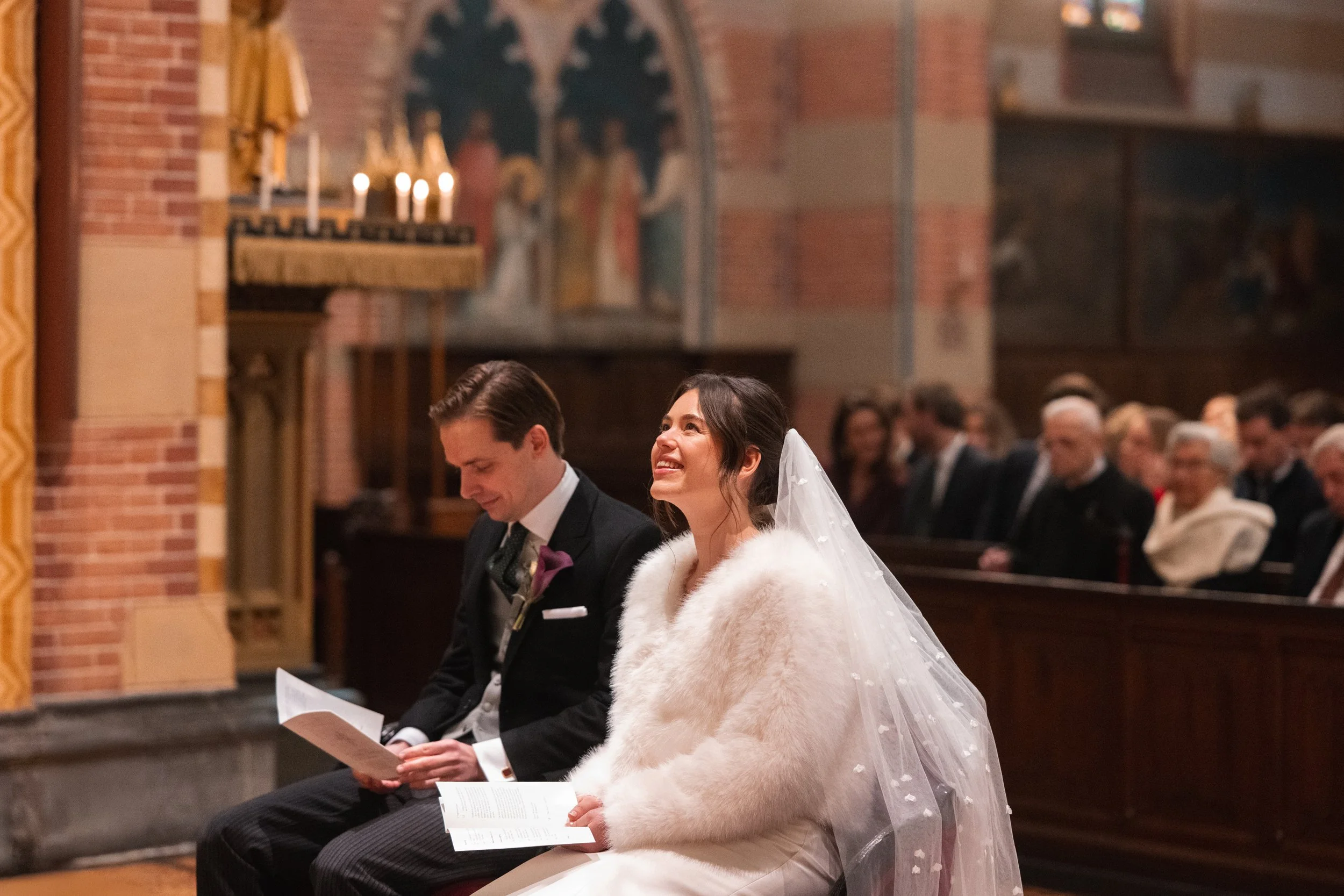 Bride and groom sitting in church during wedding ceremony, with guests in background.
