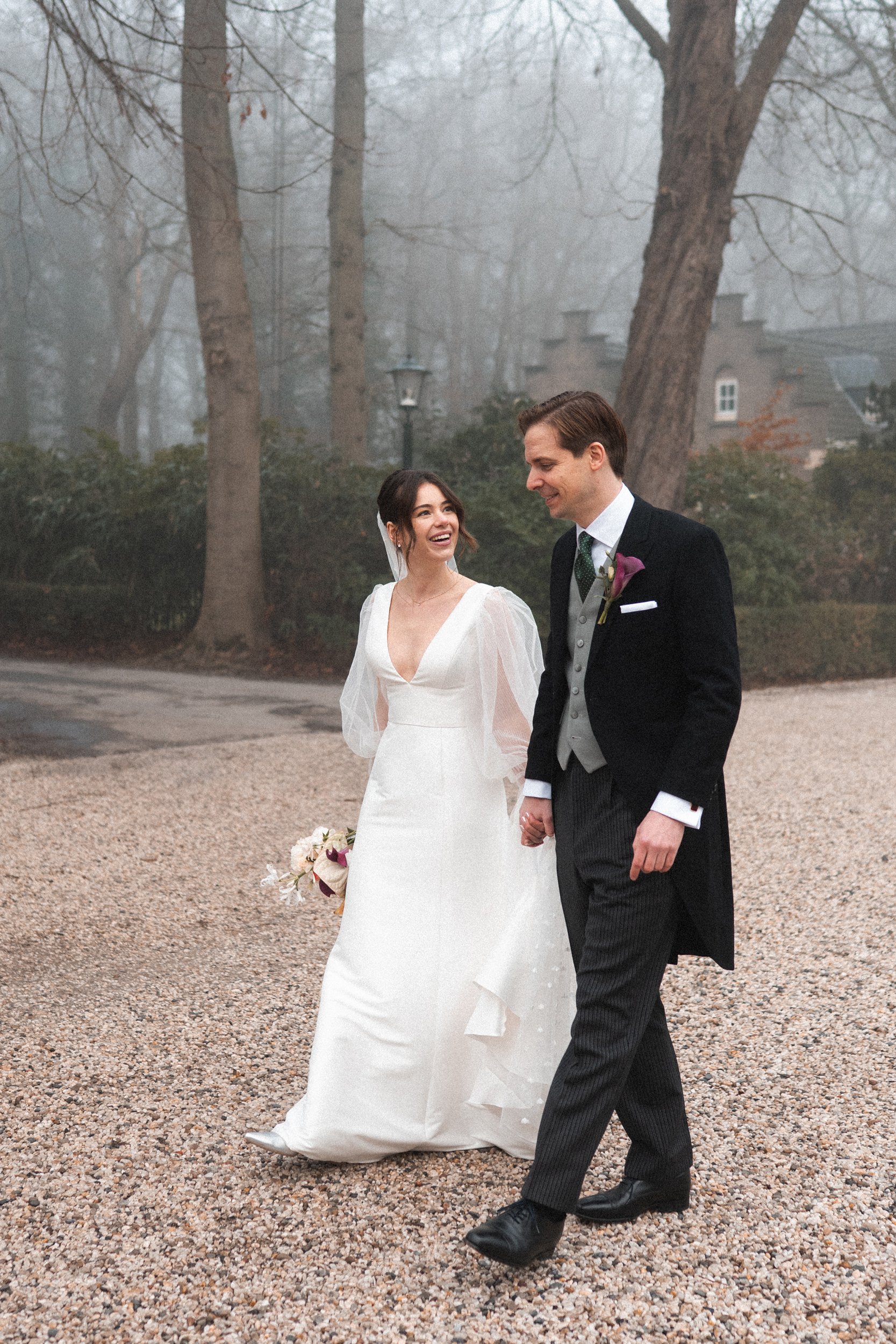 A bride and groom walking hand in hand outdoors on a foggy day, smiling warmly at each other, surrounded by tall trees and a gravel path.