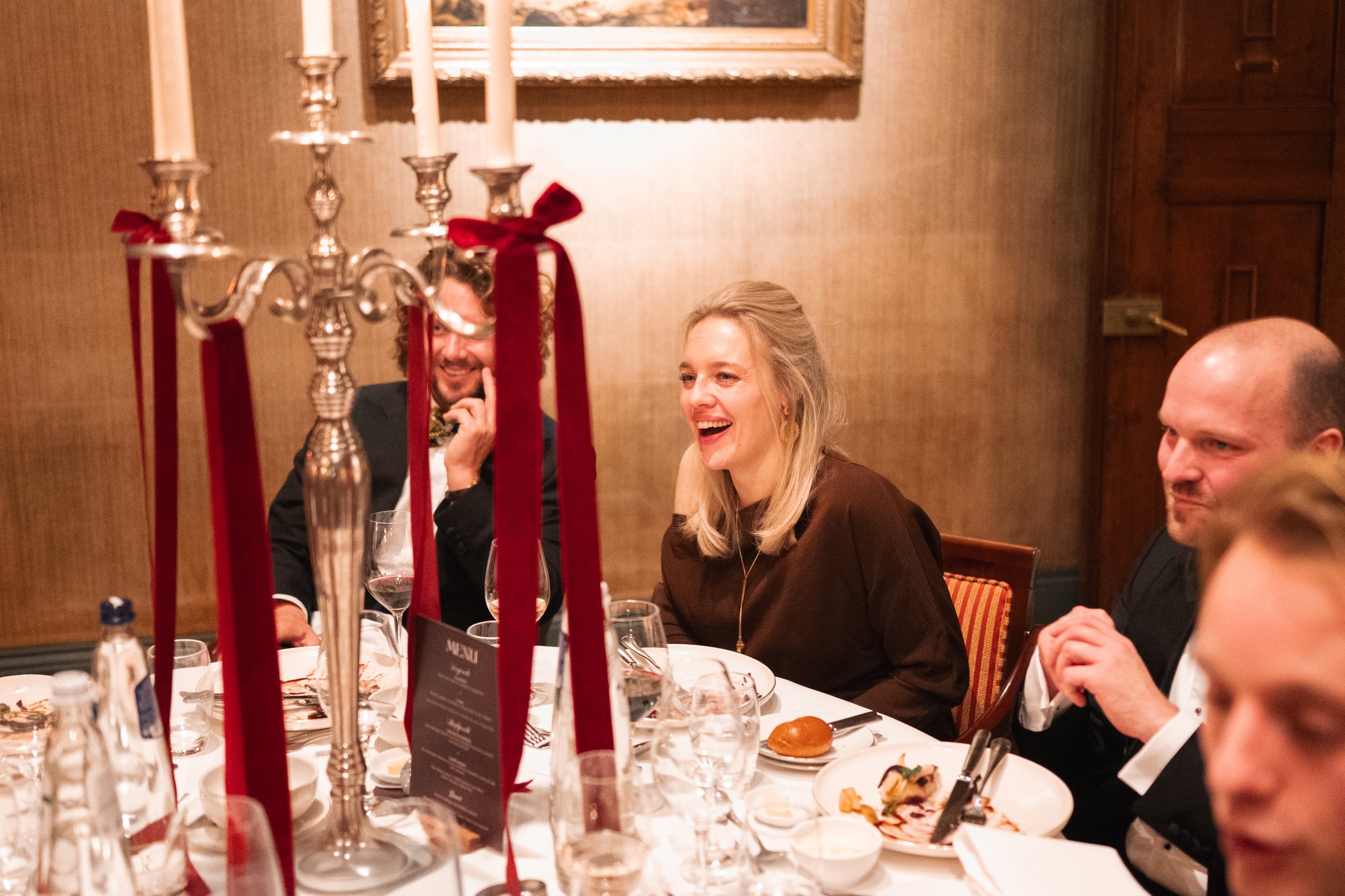 People sitting at a formal dinner table enjoying conversation, with a large silver candelabrum with red ribbons in the foreground, in a warmly lit room.