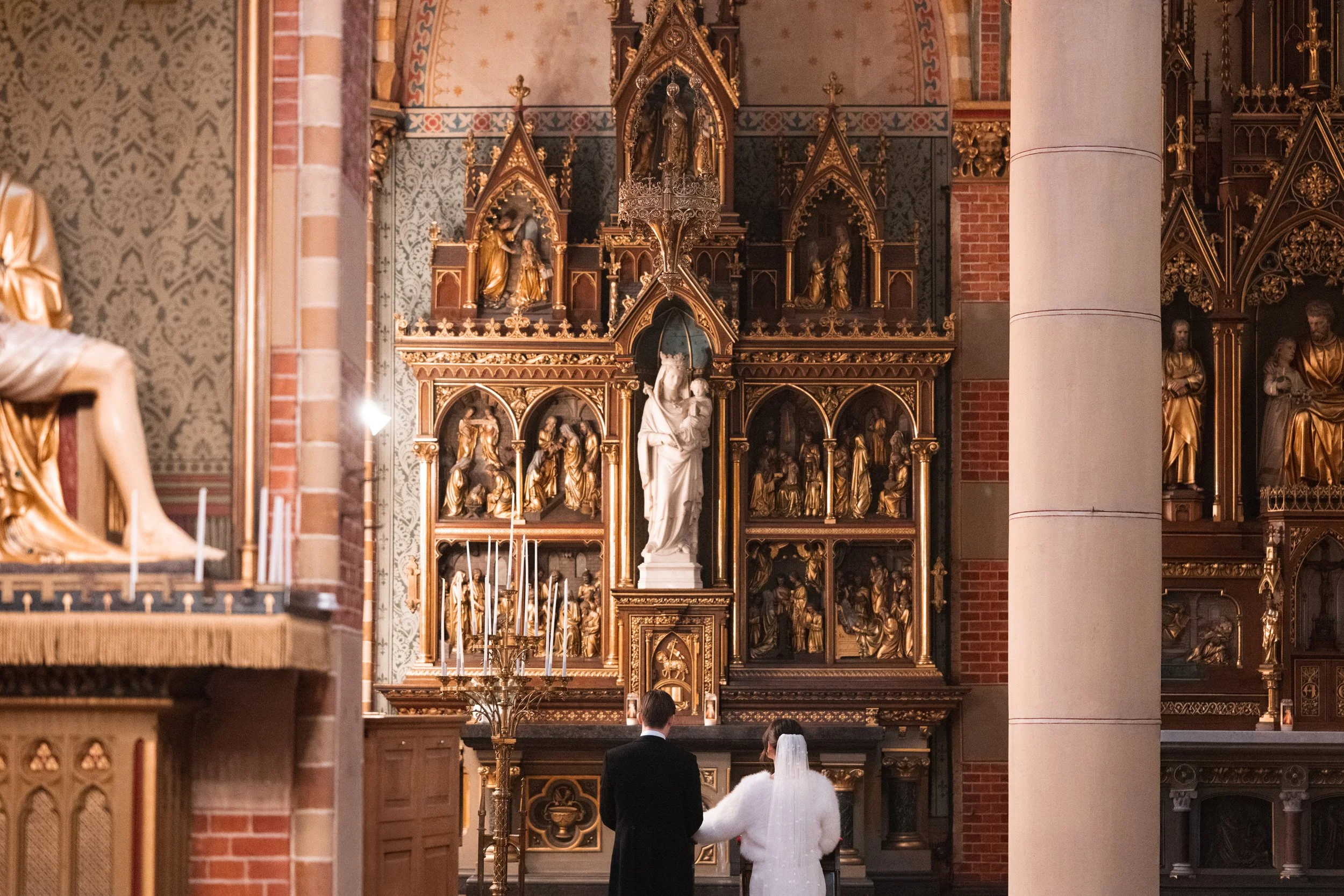 A bride and groom standing after a wedding ceremony inside a church, facing an ornate altar with religious statues and gold embellishments.