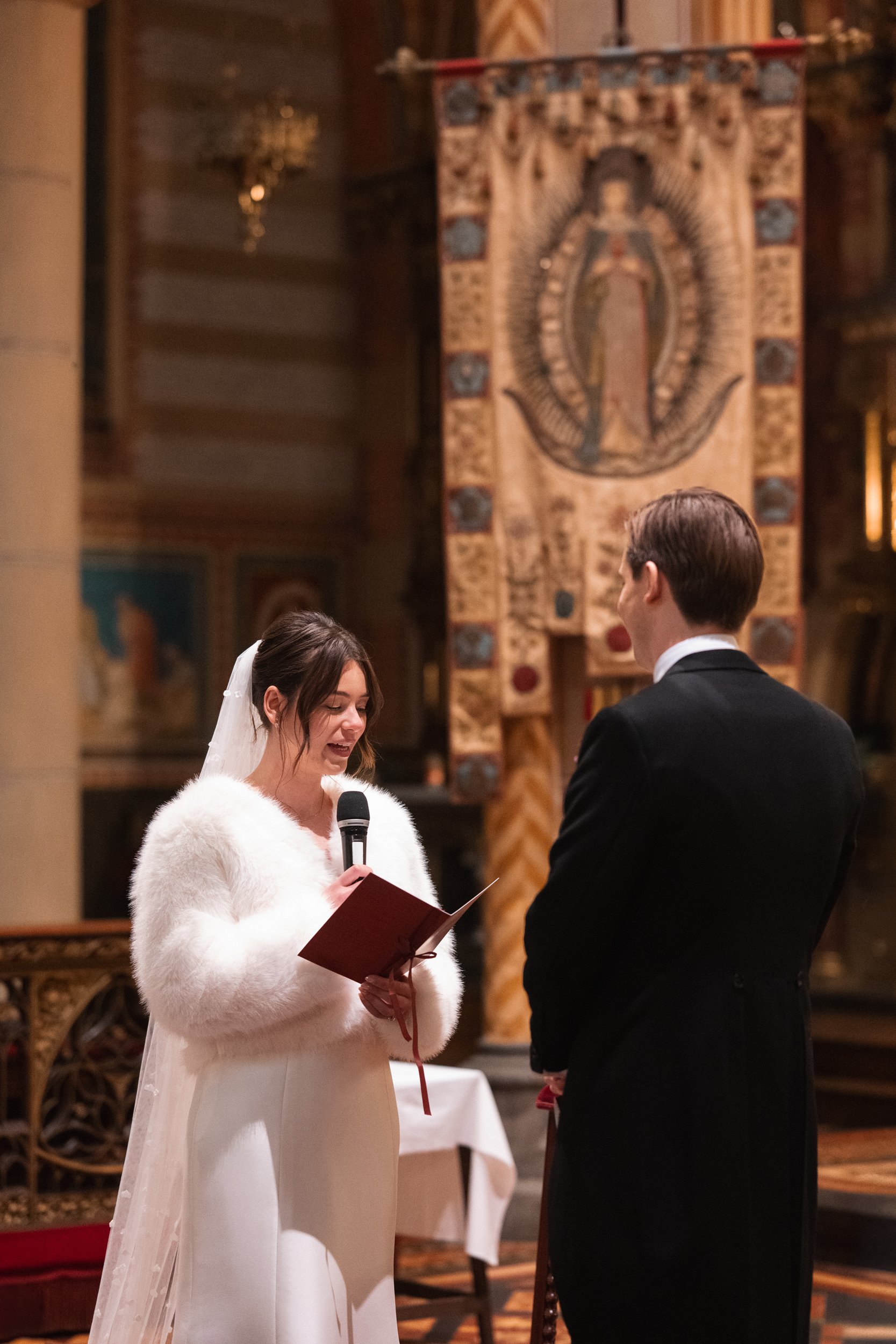 A bride holding a microphone and reading from a red book during her wedding ceremony, standing in front of a groom in a black suit in a church with ornate decorations and a religious tapestry in the background.