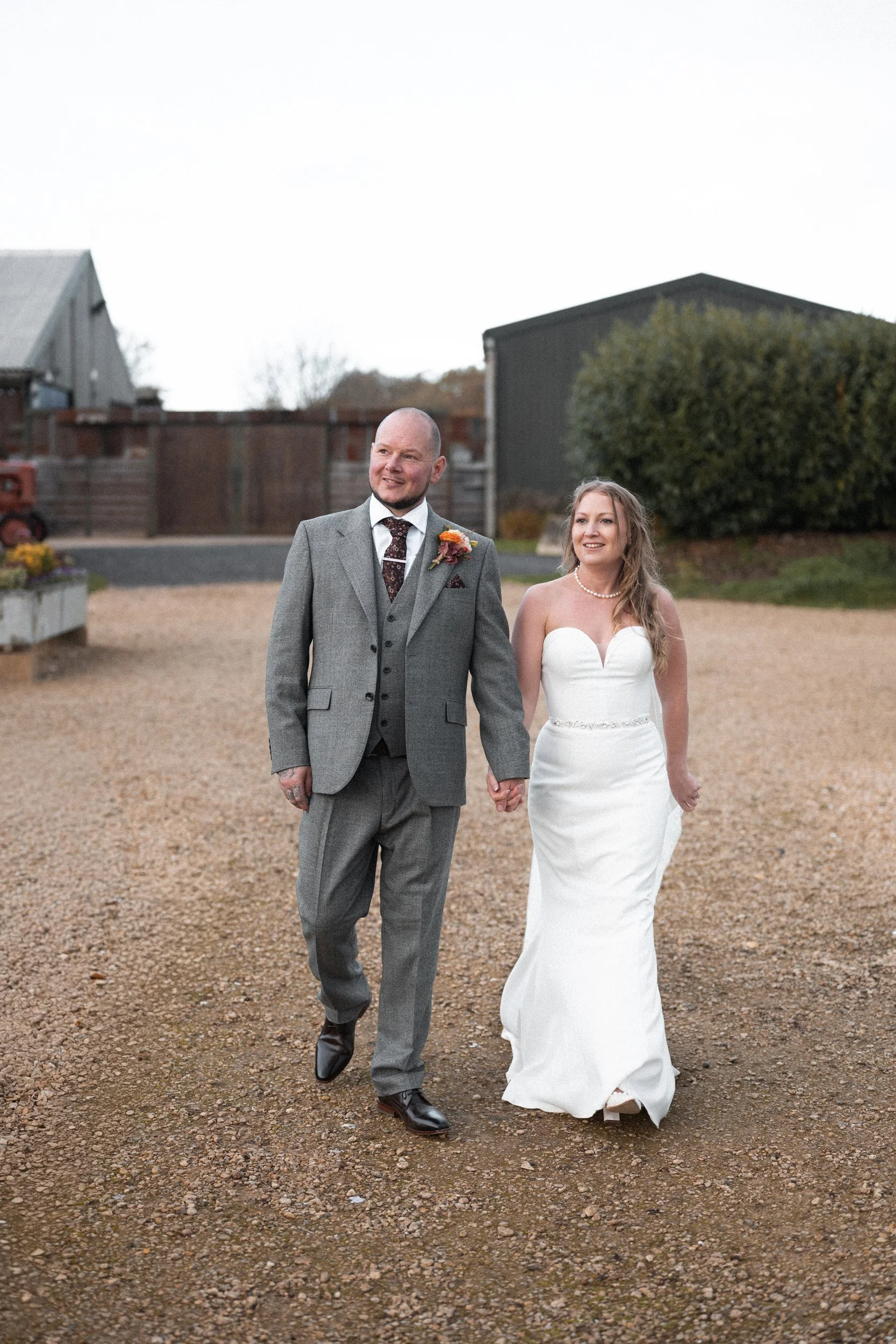 A newly married couple holding hands and walking outdoors on a gravel path, with a bride in a white strapless wedding gown and a groom in a gray suit, both smiling.