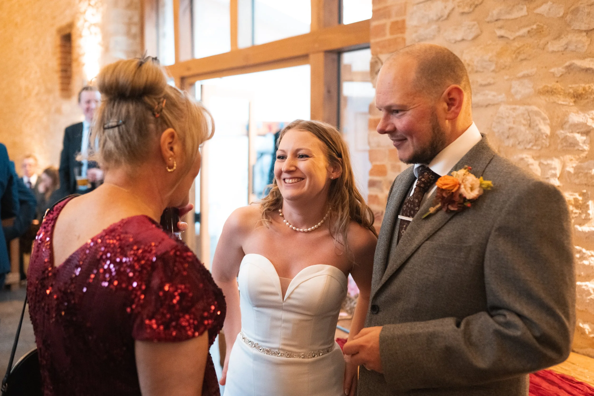 A smiling bride in a white wedding dress talking with an older woman and a man in a suit at a wedding reception inside a venue with brick walls.