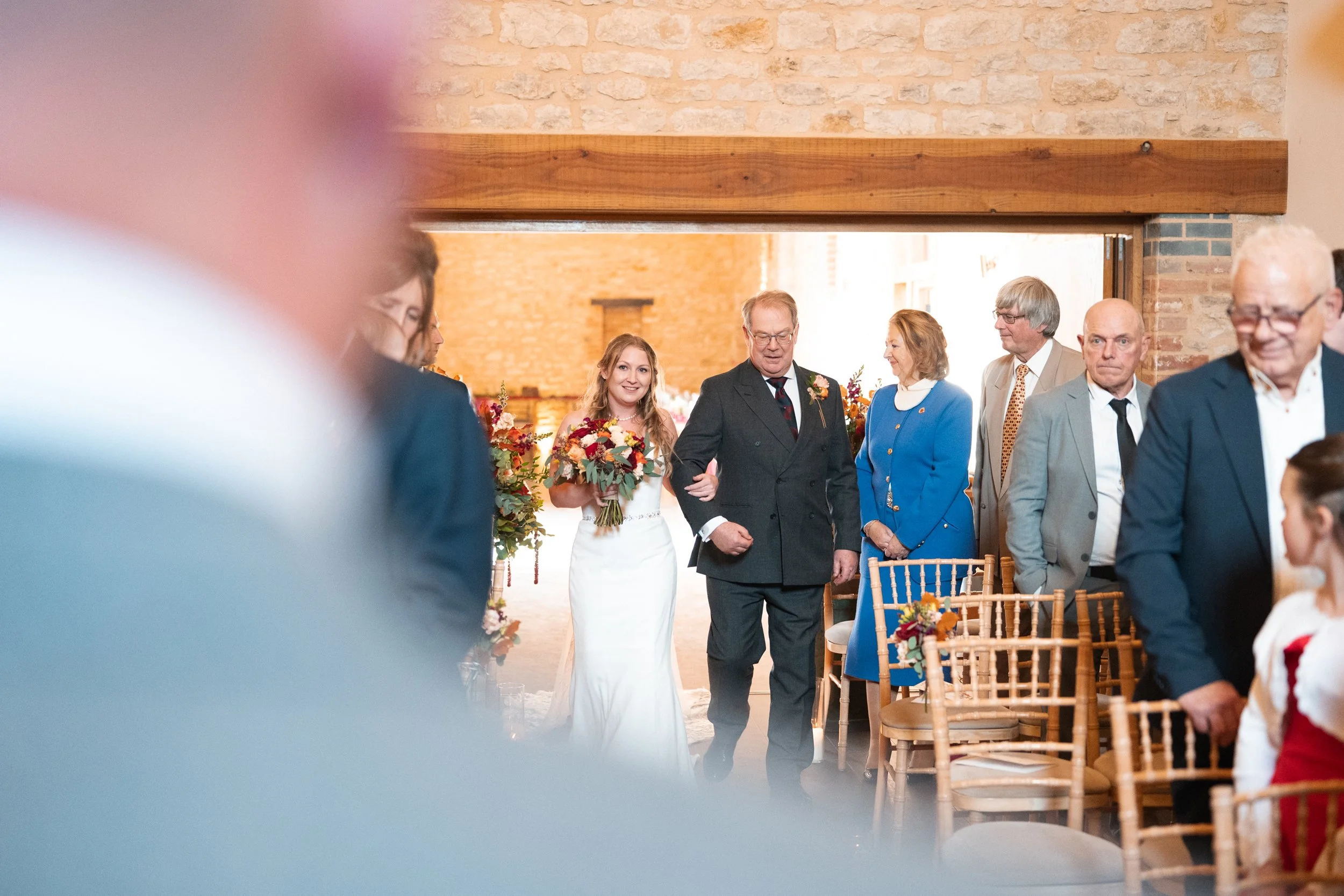 Bride walking down the aisle with her father at a wedding ceremony, surrounded by guests seated in chairs, with a rustic brick interior and floral decorations.