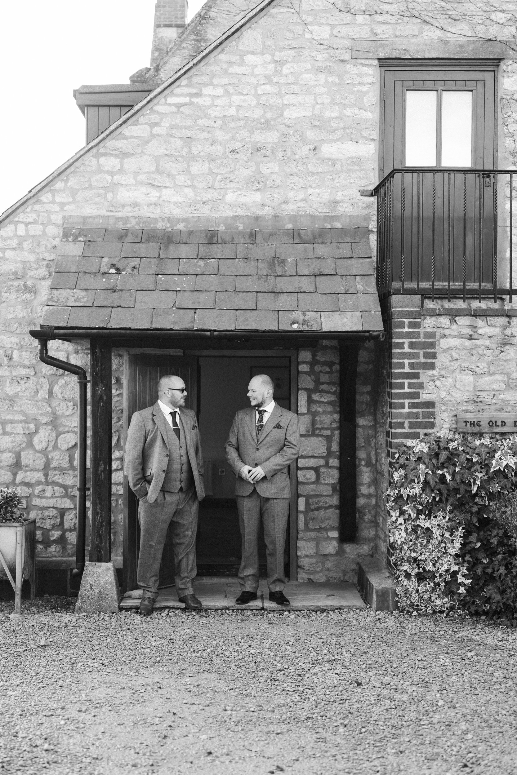 Two men in suits stand and talk in front of a stone building with a small porch, balcony, and a window.