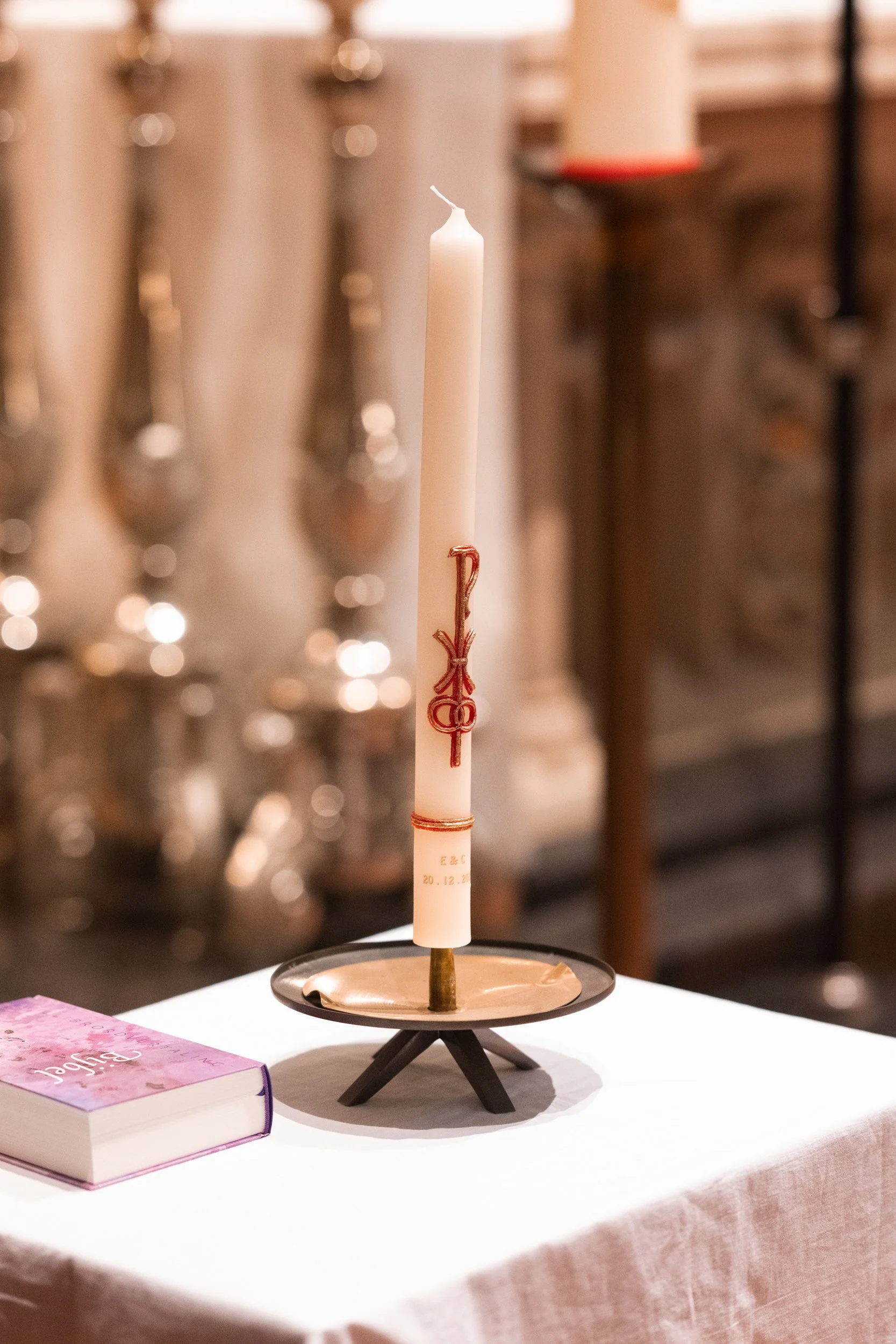 A white candle with religious symbols and words, placed in a holder on a table next to a pink and purple prayer book.