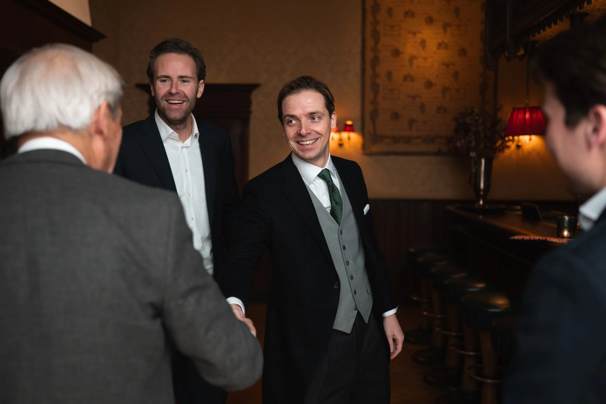 A group of four men dressed in formal attire, shaking hands and engaging in conversation in a warmly lit room with vintage decor.