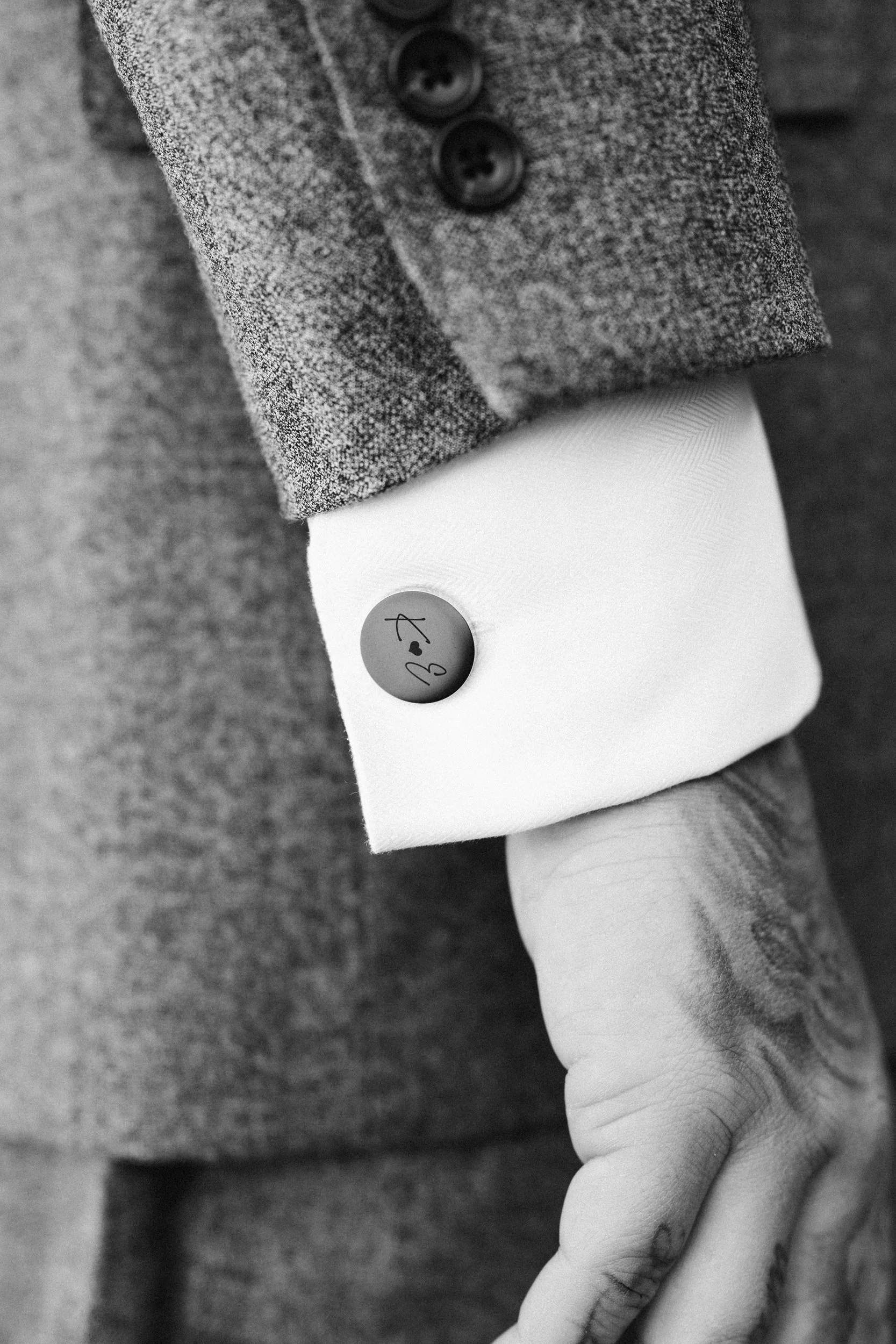Close-up of a person's wrist with a white shirt cuff and a button featuring a signature or logo, and a gray tweed jacket sleeve with black buttons.