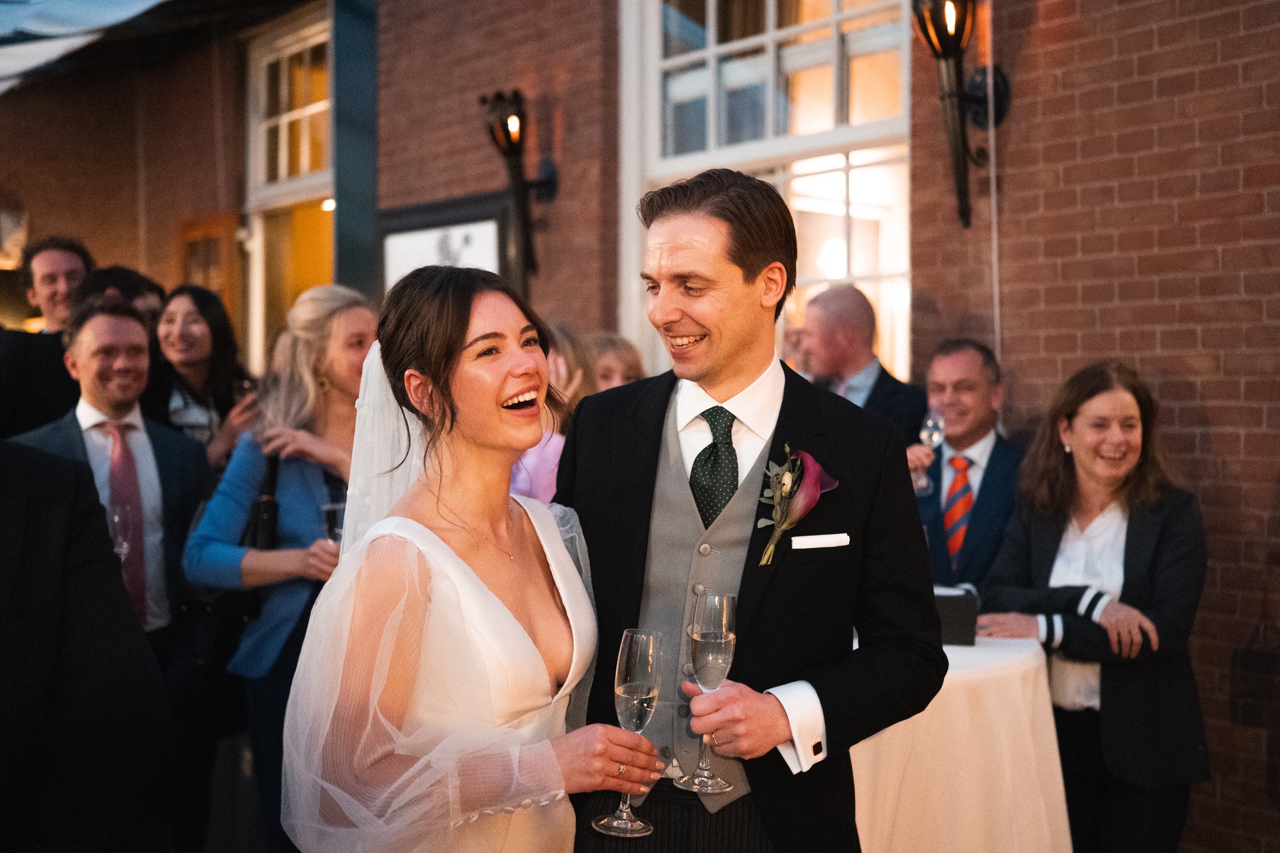A bride and groom smiling and holding champagne glasses at their wedding reception, surrounded by guests.