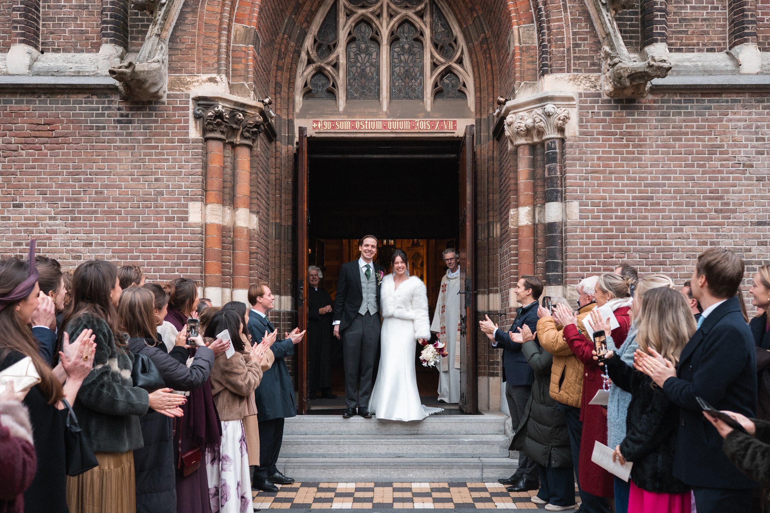 A bride and groom standing at the entrance of a church, smiling, surrounded by guests clapping and taking photos during a wedding celebration.