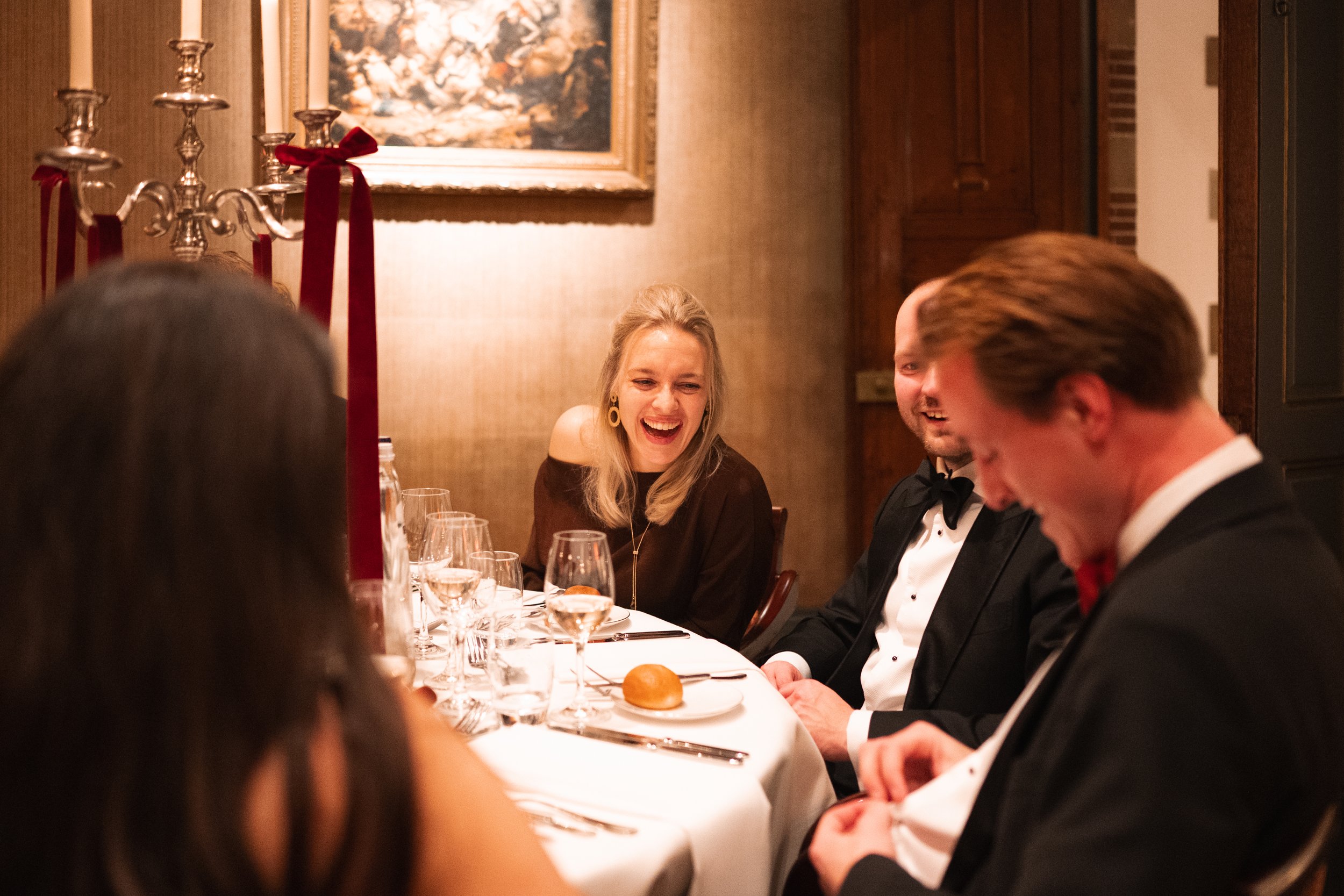 Group of adults dressed in formal attire enjoying a dinner party at a restaurant or banquet hall, with a woman in black laughing and others smiling and engaged in conversation.